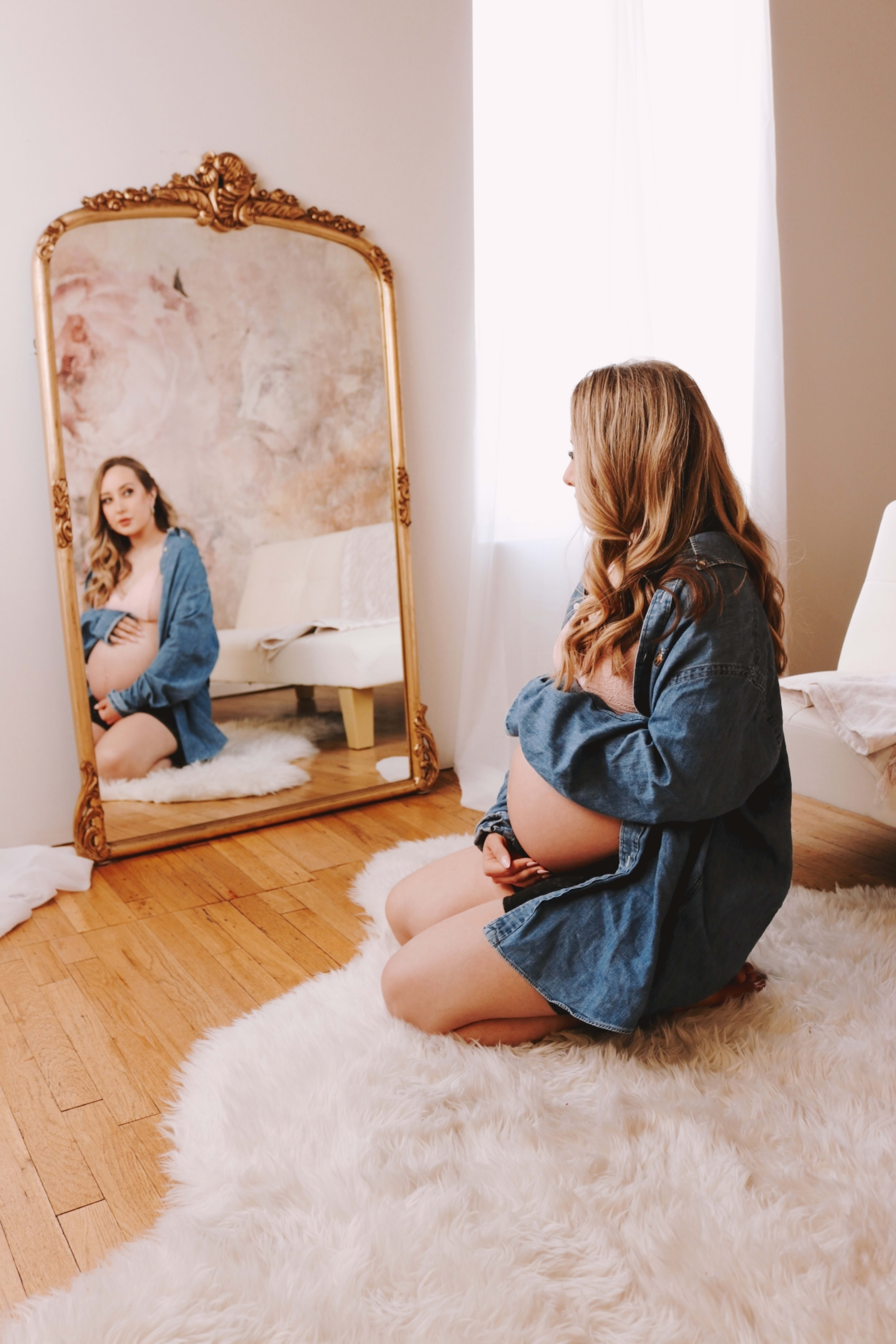 Une femme enceinte assise sur un sol blanc lors d'une shooting photo de maternité devant un miroir.