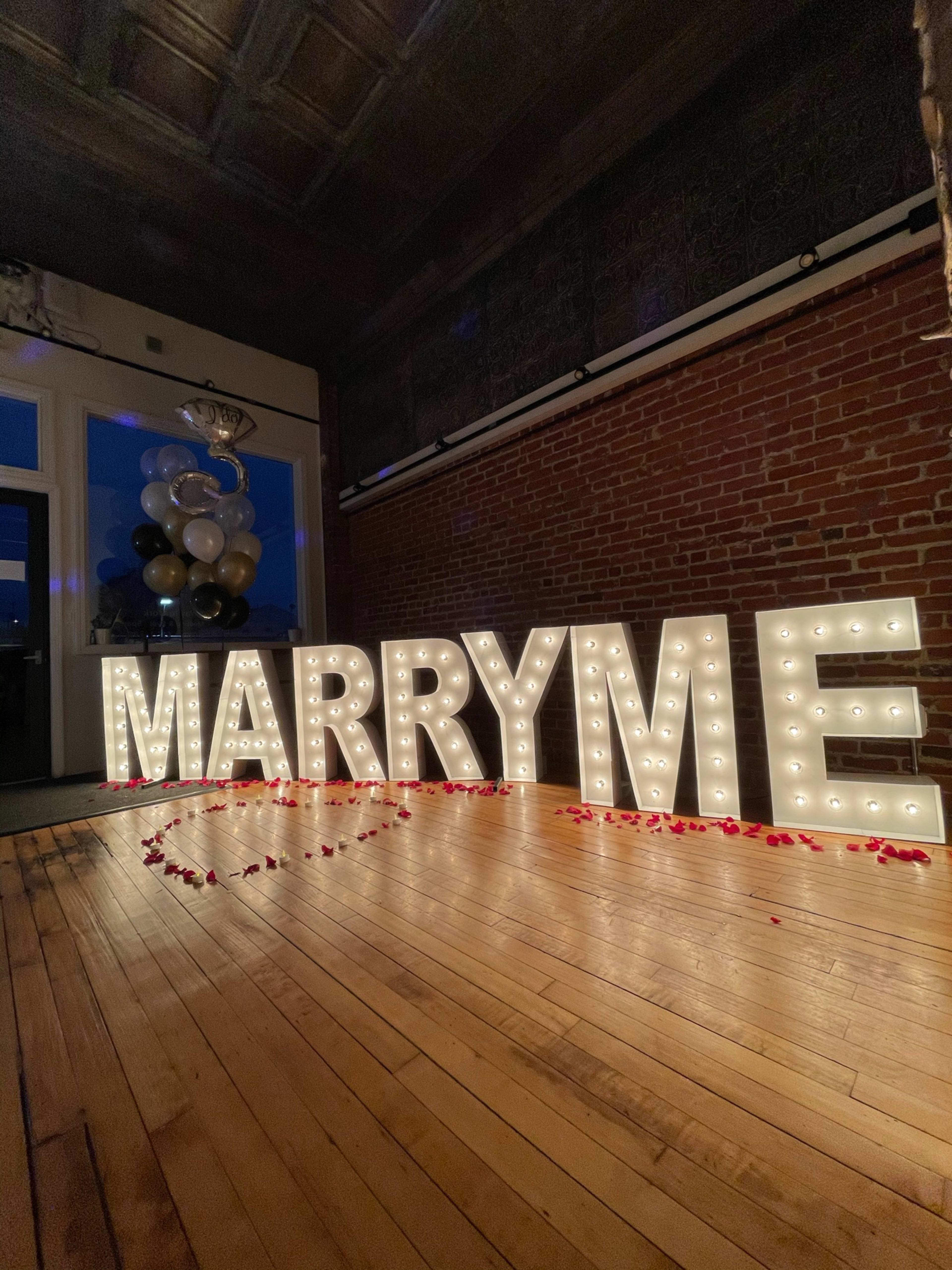 A beige marquee sign that says marry me on a wooden floor at an engagement party.