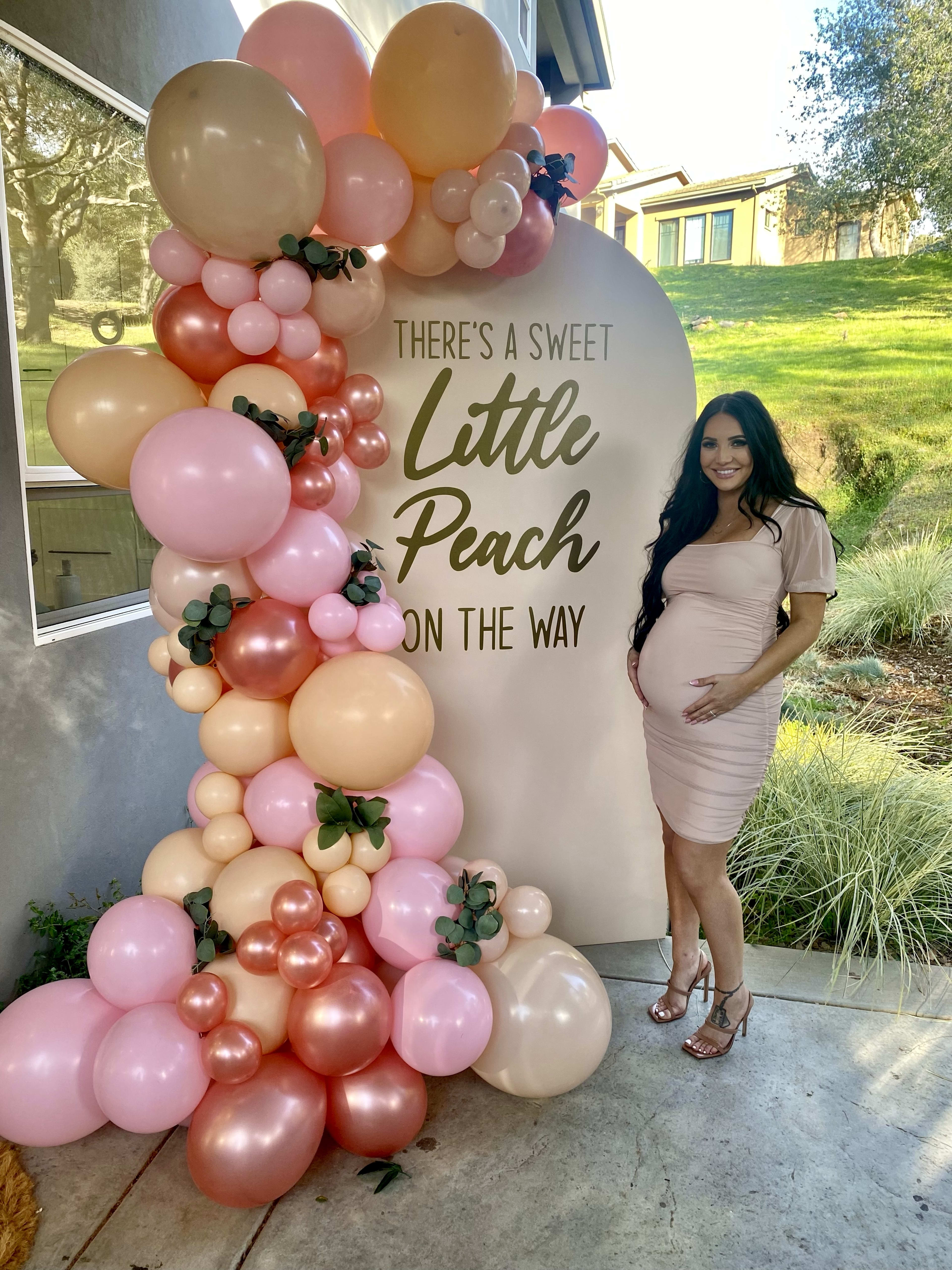 A pregnant woman standing in front of an outdoor baby shower sign with pink balloons.