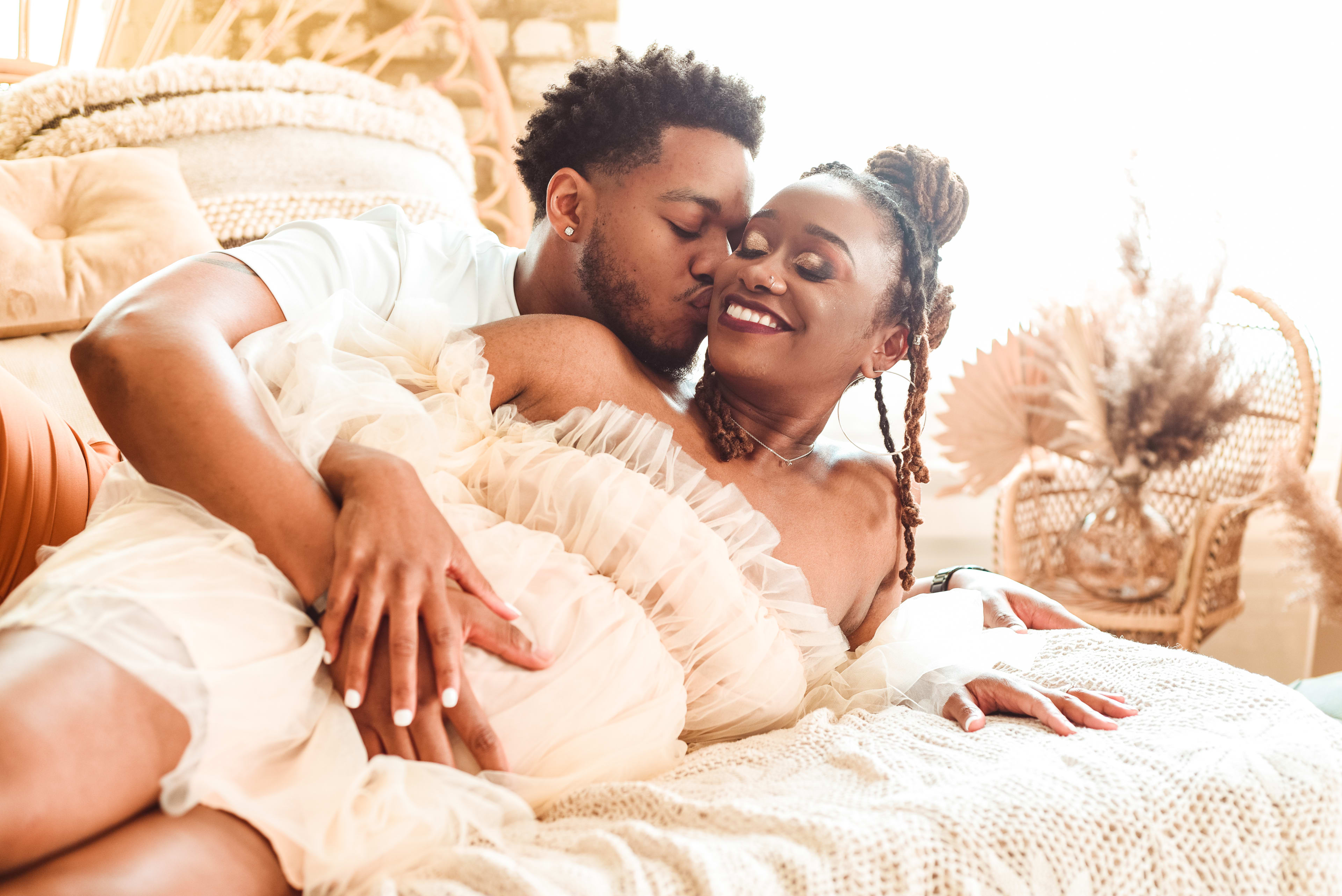 A white maternity photo shoot of a man and woman lying on a boho-style bed in beige bedding.