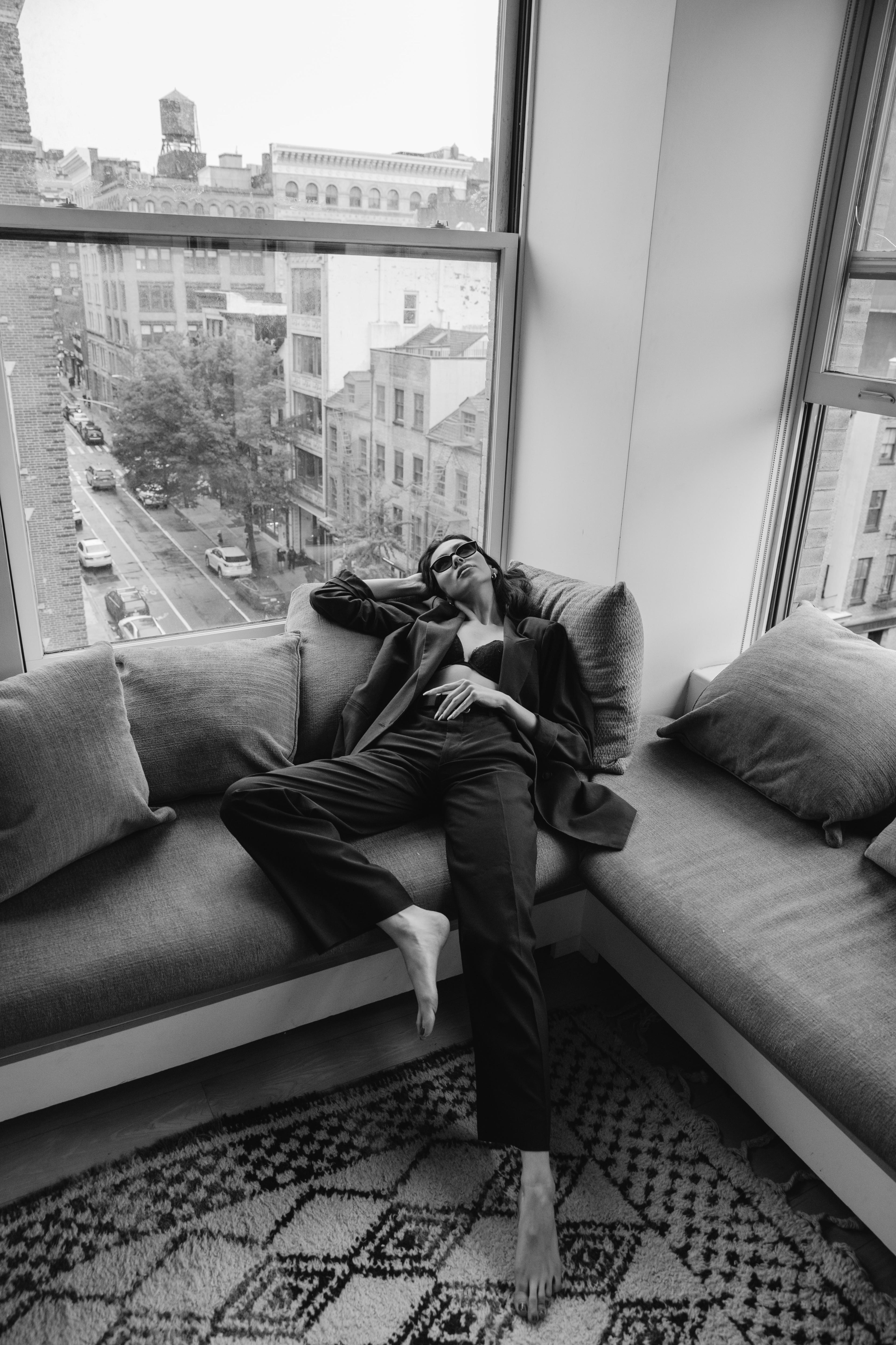 A black and white photoshoot of a person laying on a couch in front of a window.