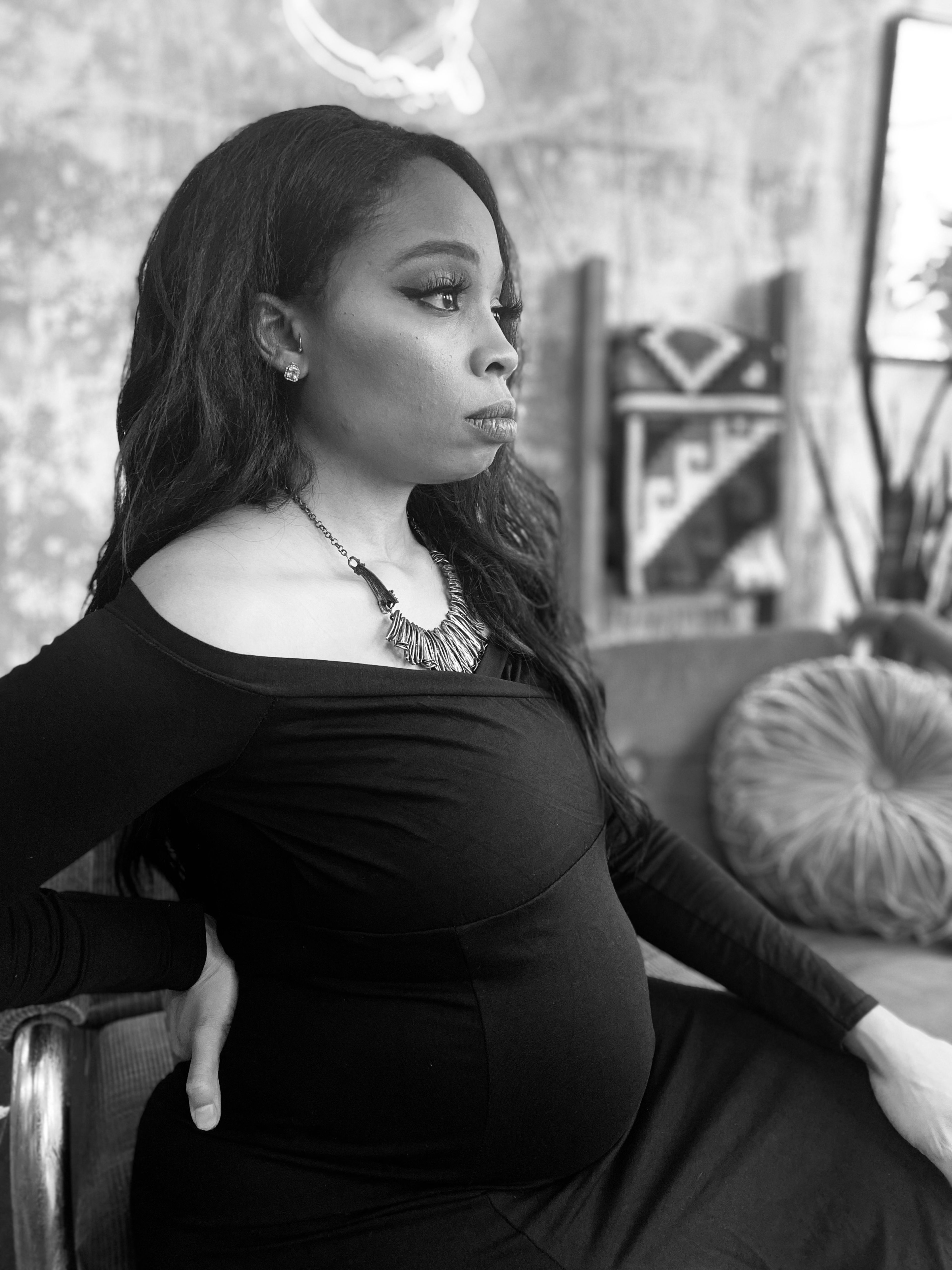 A maternity photoshoot of a woman sitting in a black chair, captured in black and white.