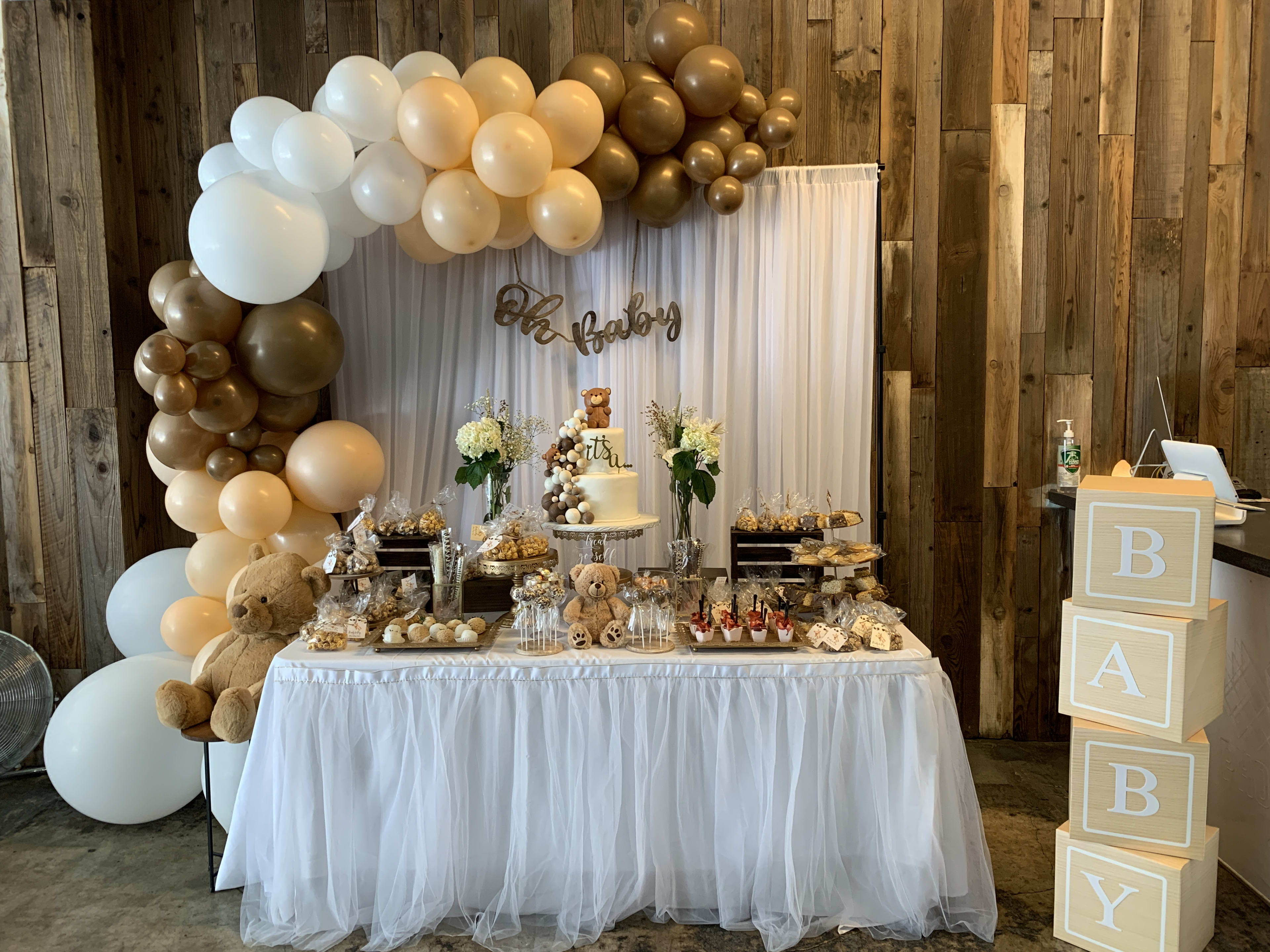 A gender-neutral baby shower table adorned with a teddy bear cake and white and brown balloons.