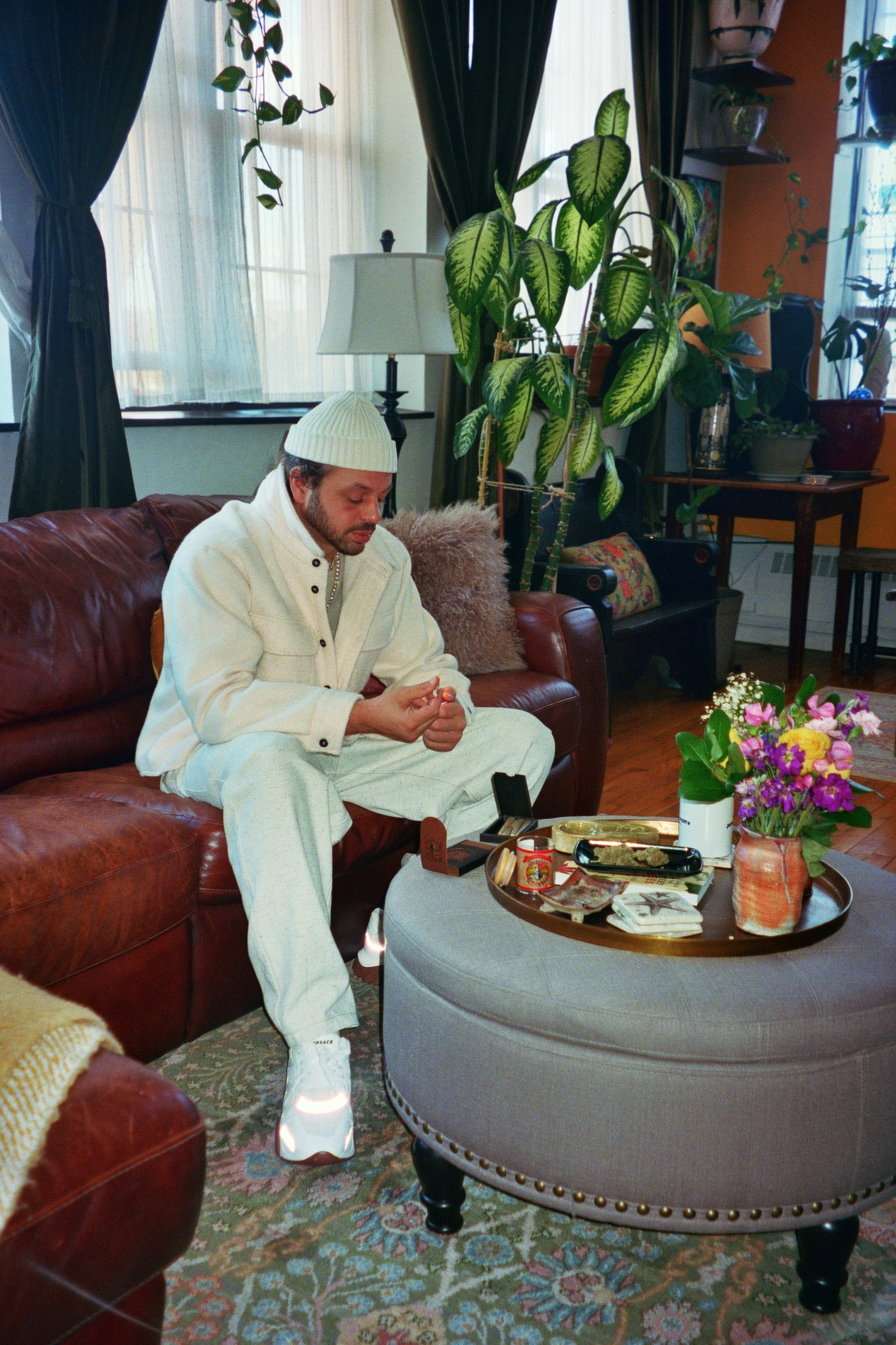 A man wearing all white sitting on a leather couch in a living room for a photoshoot.