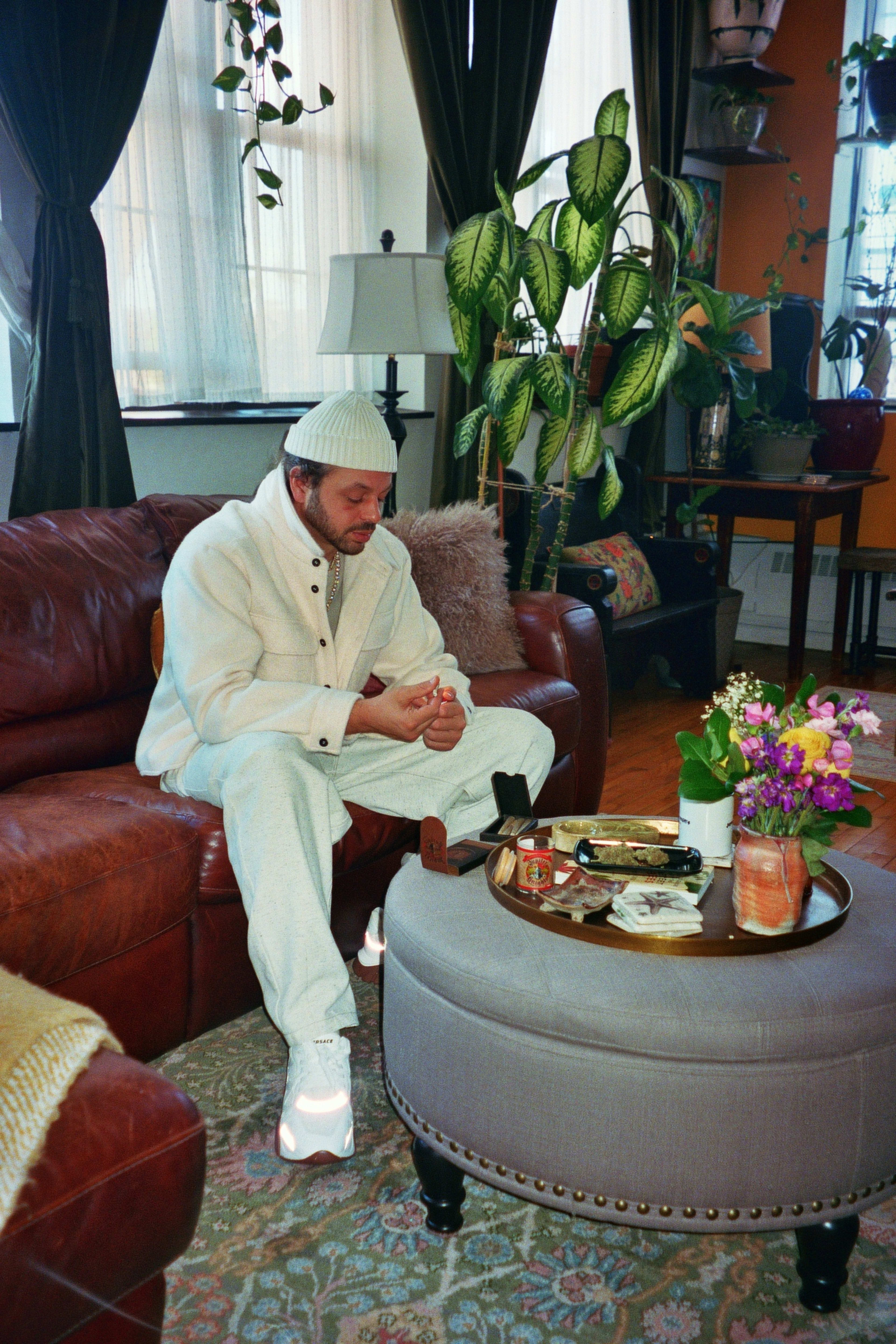 A man wearing all white sitting on a leather couch in a sitting room for a photo shoot.