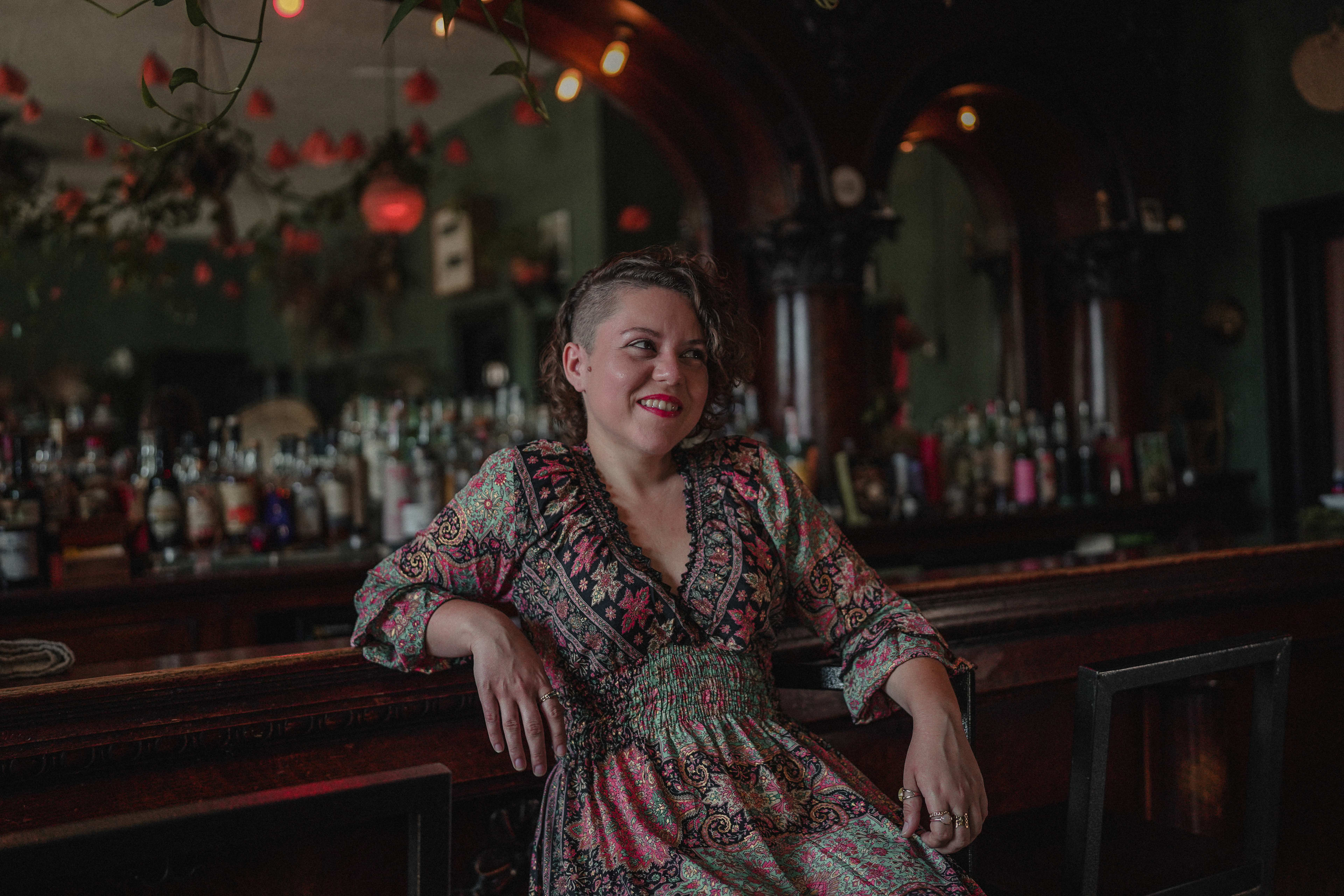 A portrait of a woman sitting at a bar with a lot of bottles behind her.