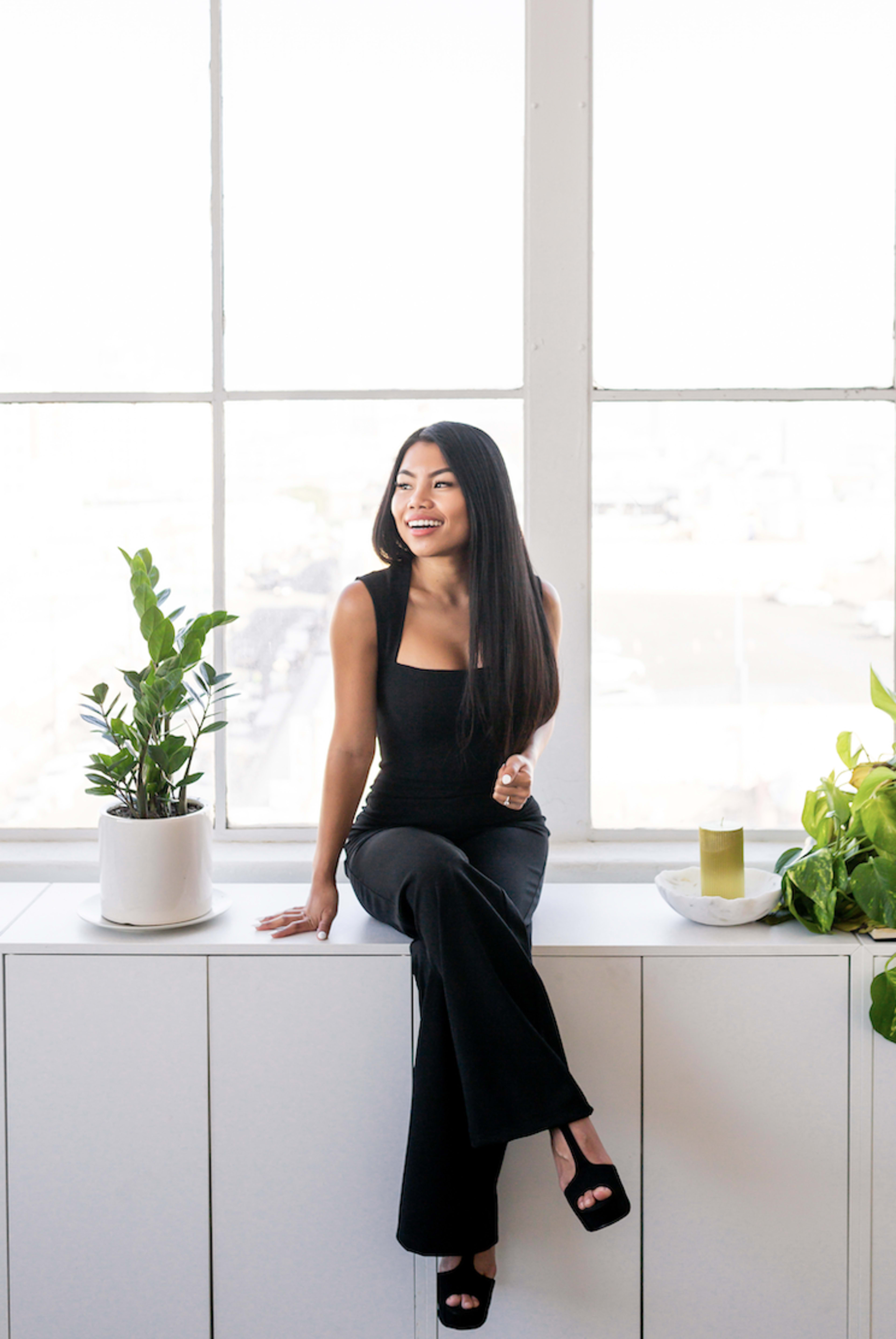 A woman posing for a photo shoot on a white counter in front of a window with plants.