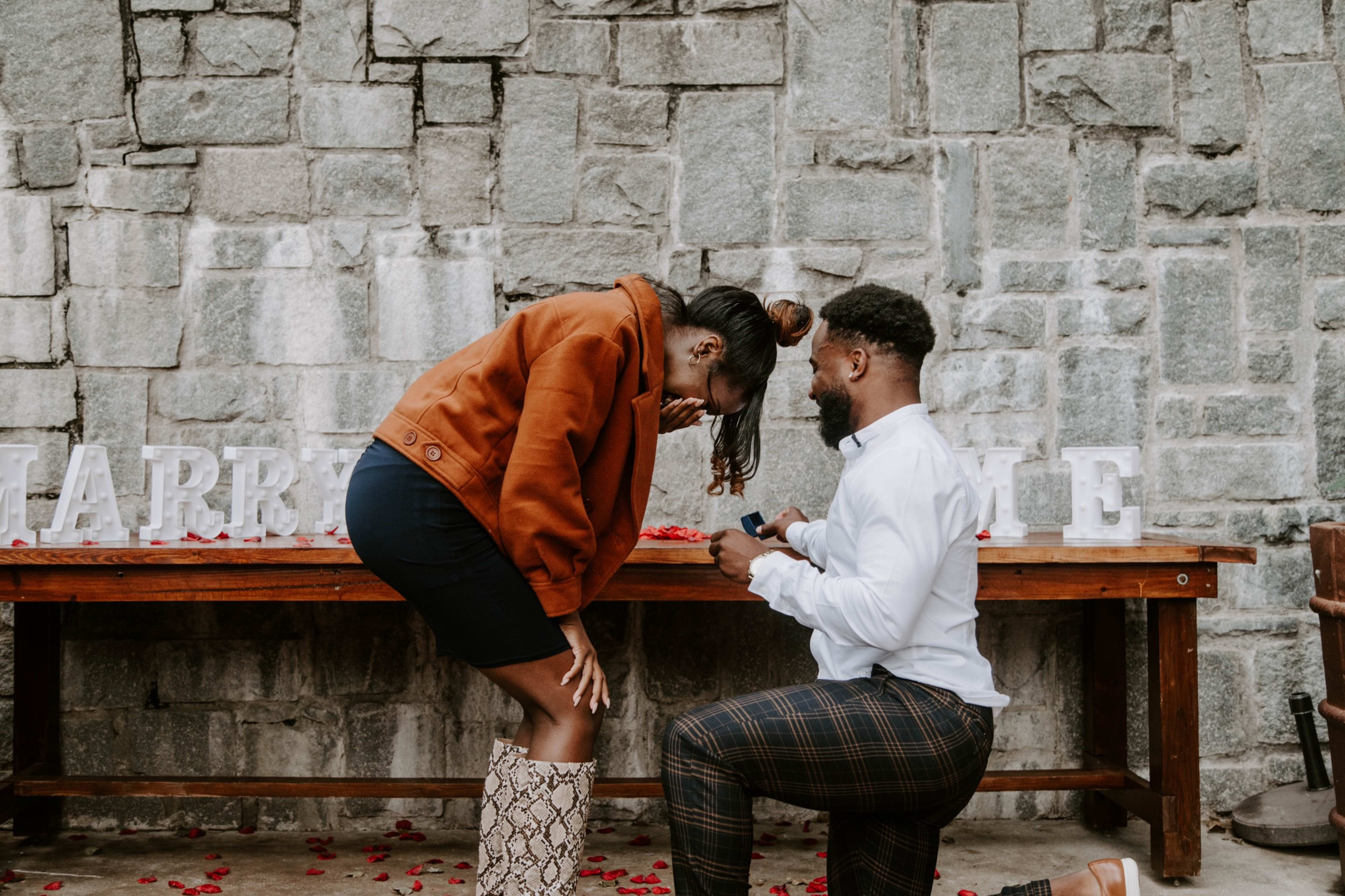 A man proposes to a woman on a white bench in a rustic setting.