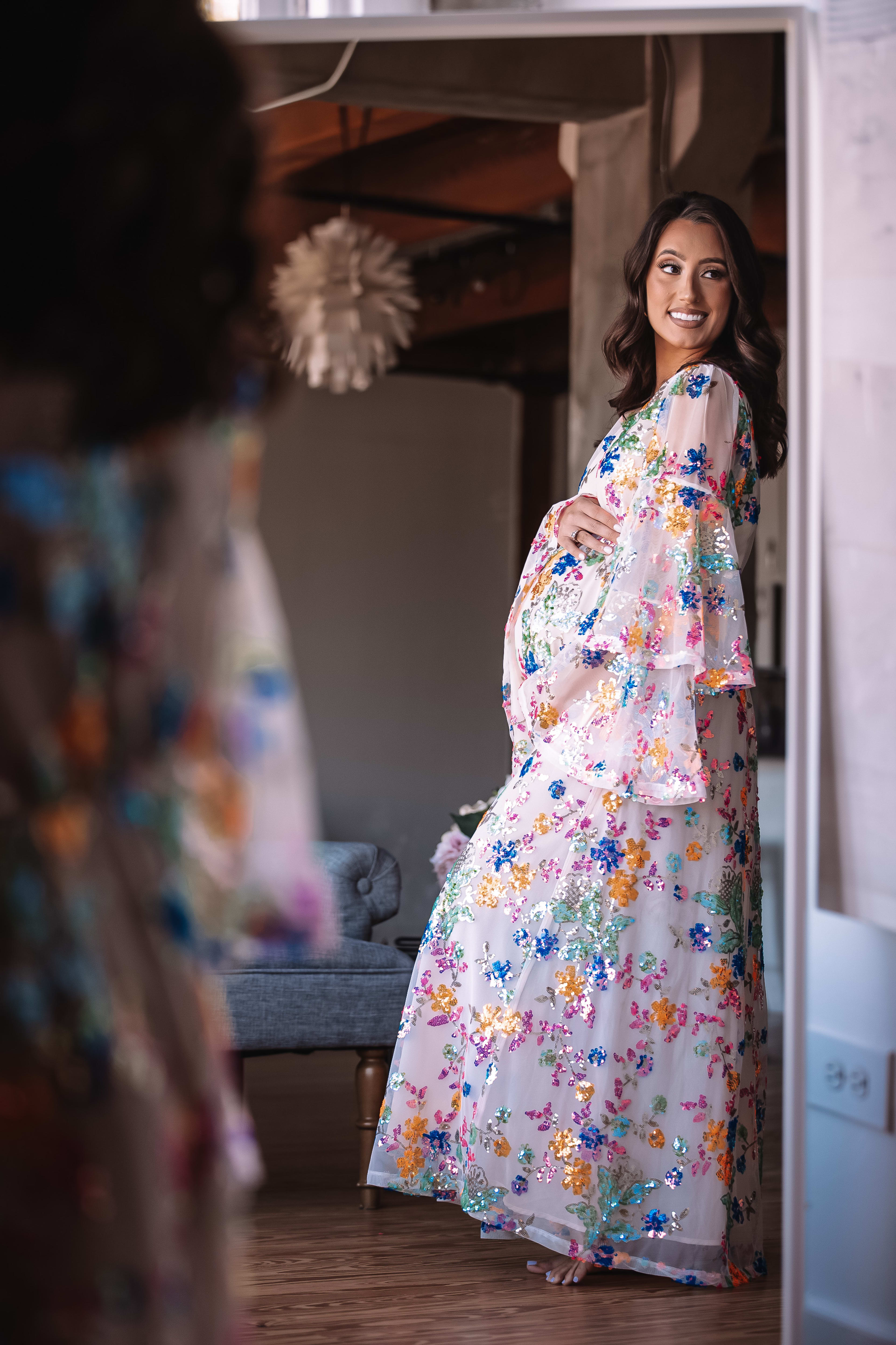 A maternity photo shoot of a pregnant woman in a floral dress standing in front of a rustic mirror.