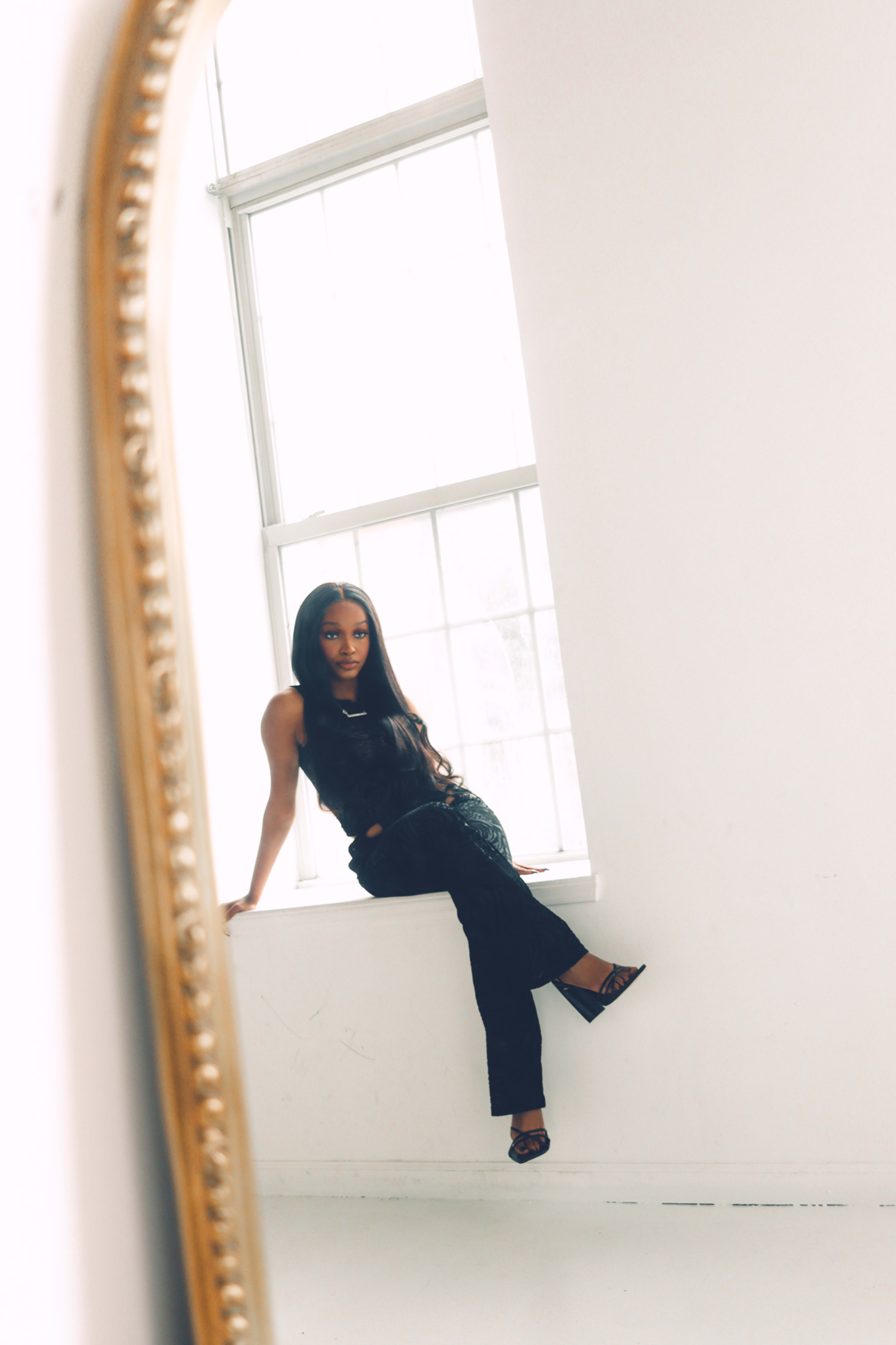 A minimalist photoshoot of a woman sitting on a black ledge in front of a white mirror.