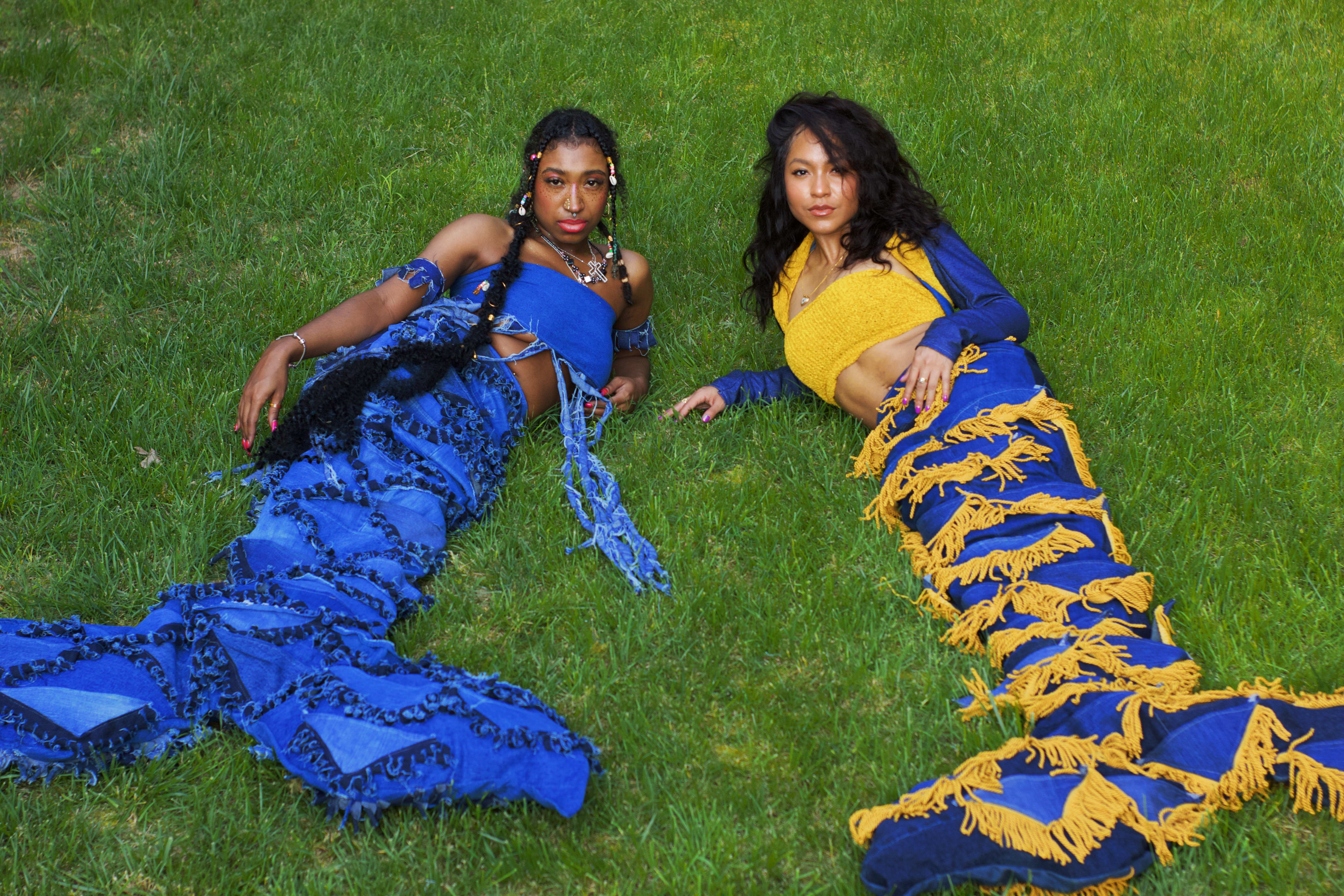 Two women in mermaid costumes in a green outdoor garden laying on the grass next to each other.