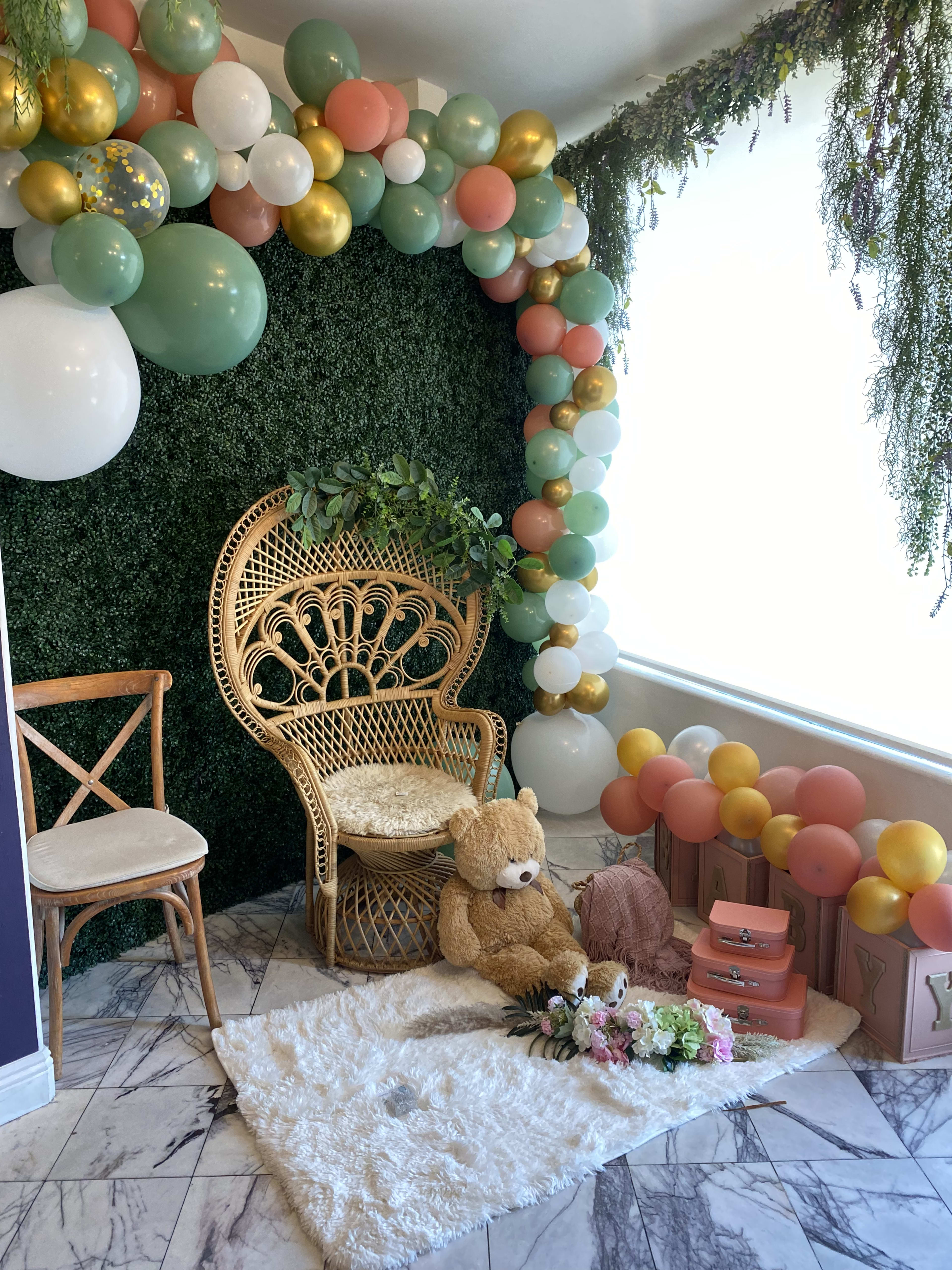 Un ours en peluche est assis devant une chaise de style boho au milieu de plantes vertes et de ballons beiges lors d'une baby shower neutre.