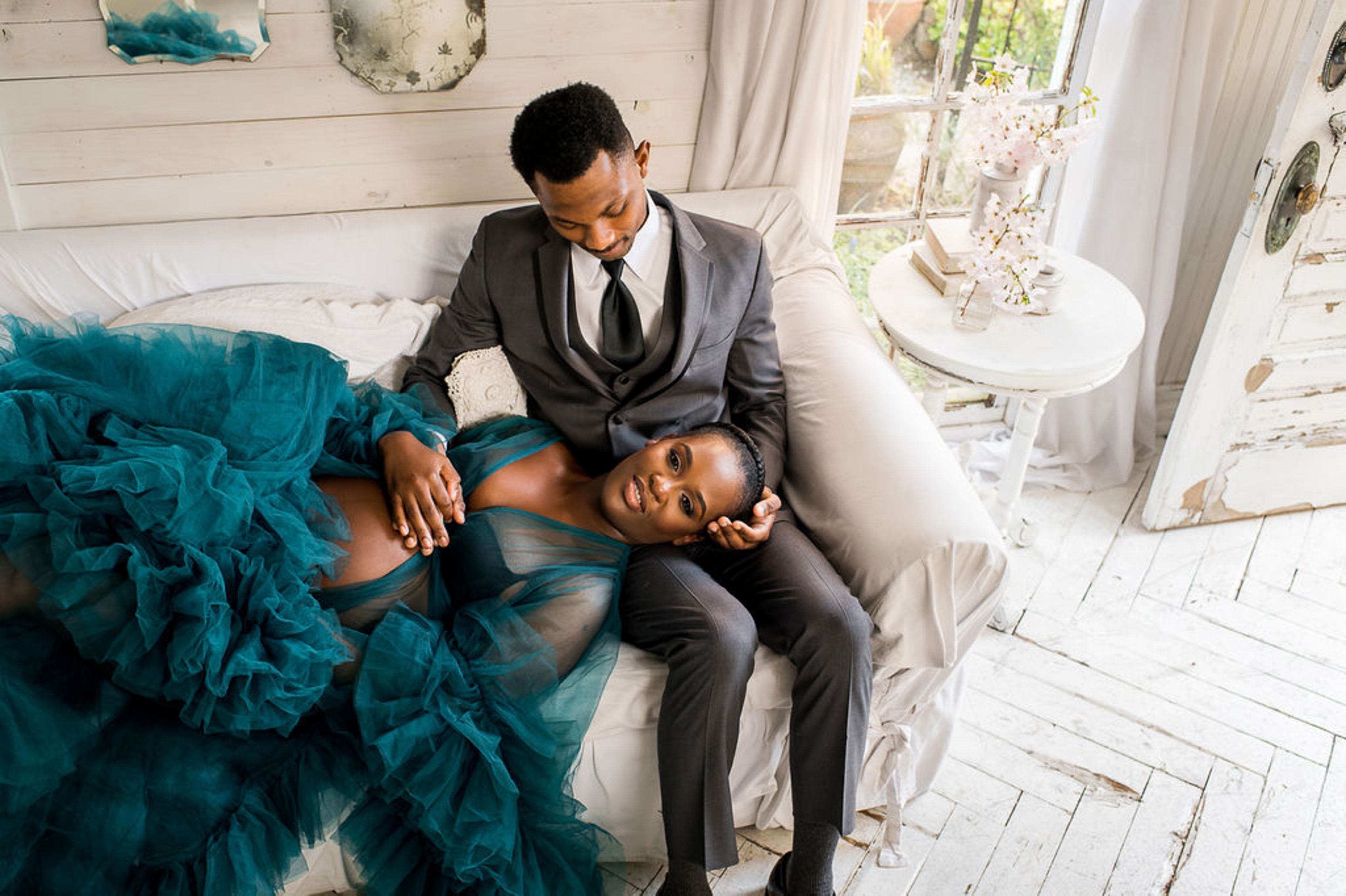 A maternity photo shoot of a man and a woman sitting on a rustic couch surrounded by blue and white accents.