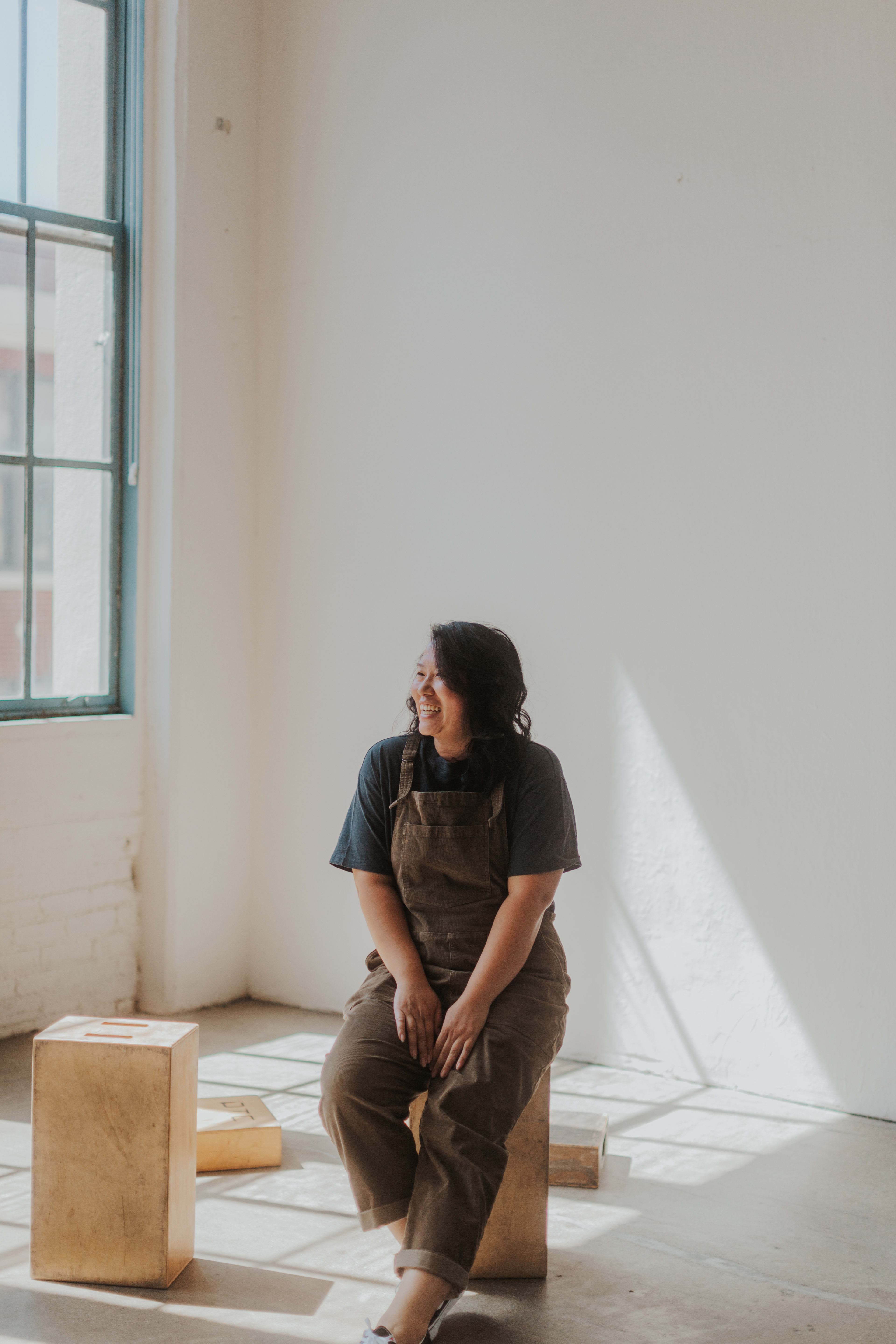 Une femme assise sur un tabouret dans un cadre minimaliste devant une fenêtre.