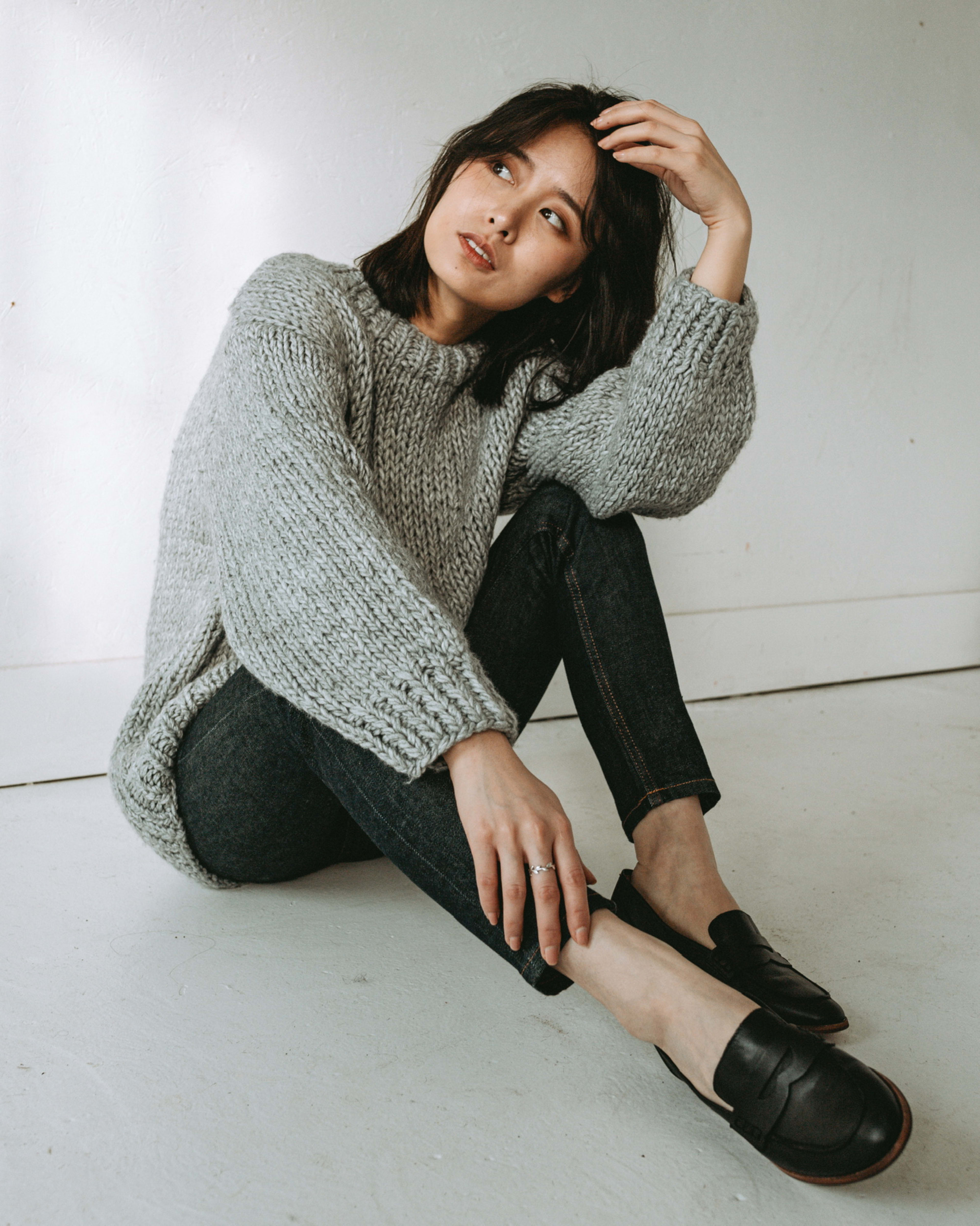 A minimalist photoshoot featuring a woman sitting cross-legged on the grey floor.