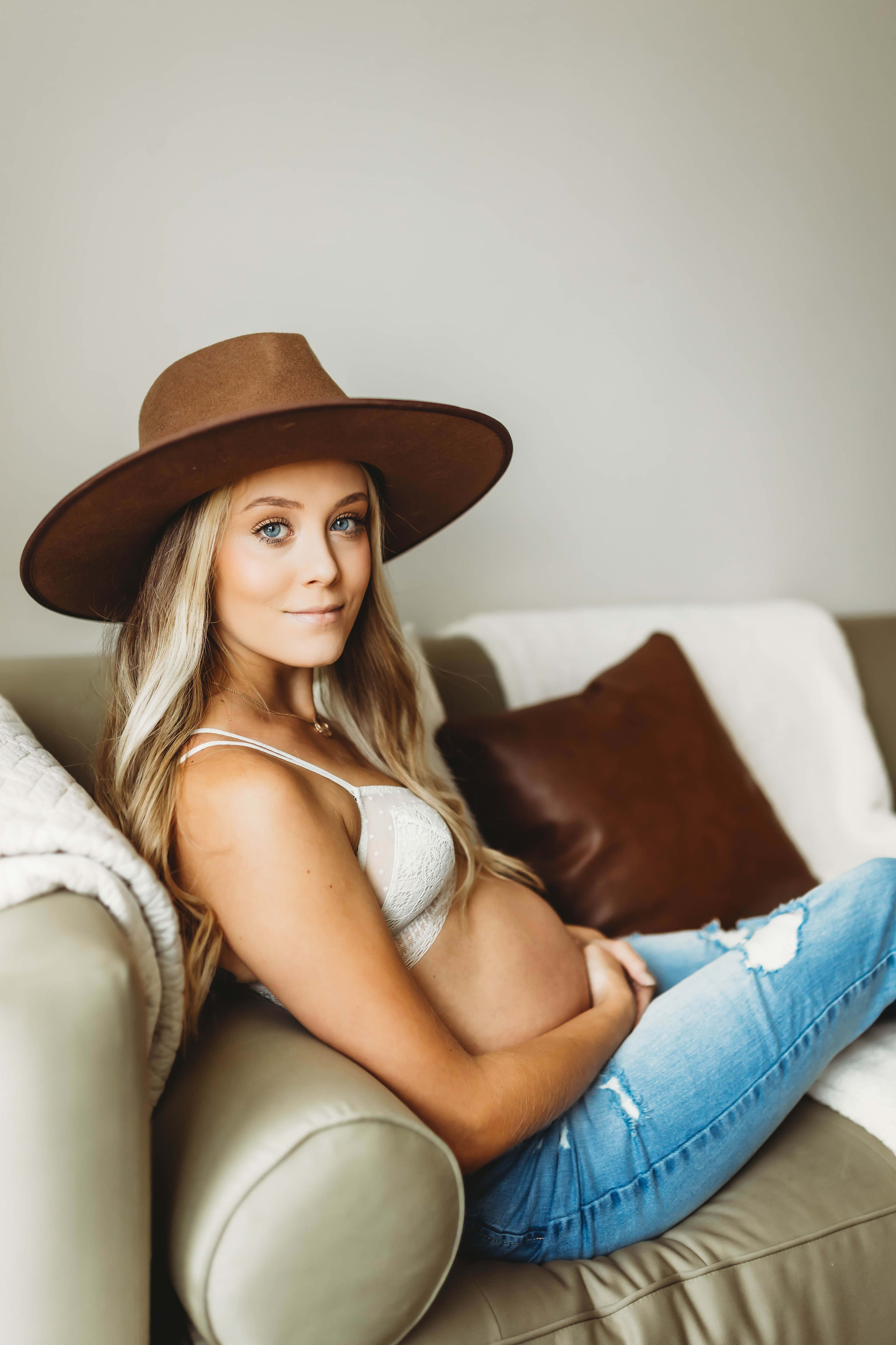 A pregnant woman wearing a brown cowboy hat sitting on a white couch during a maternity photoshoot.