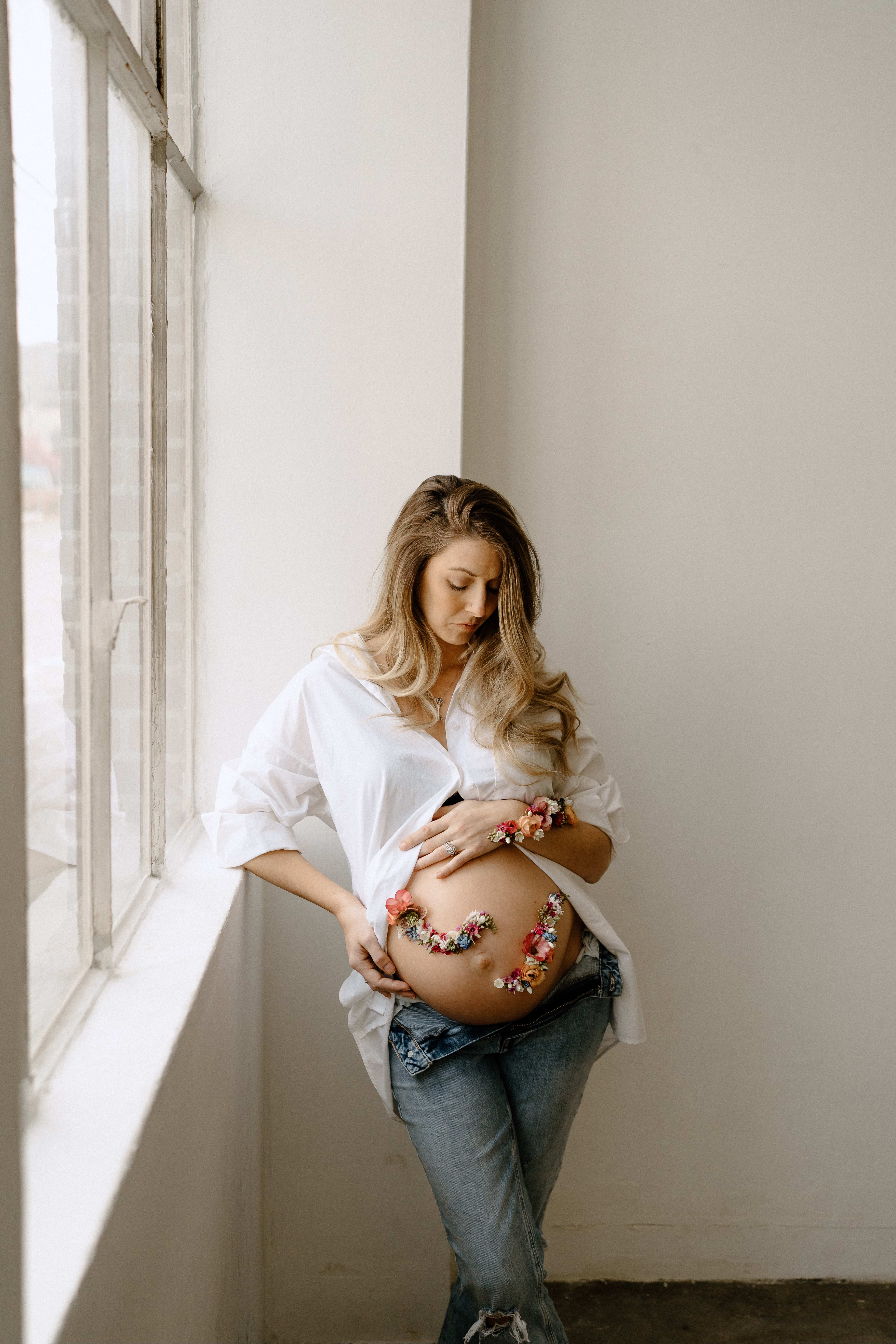 A boho-inspired maternity photo shoot featuring a white-clad pregnant woman leaning against a window sill.