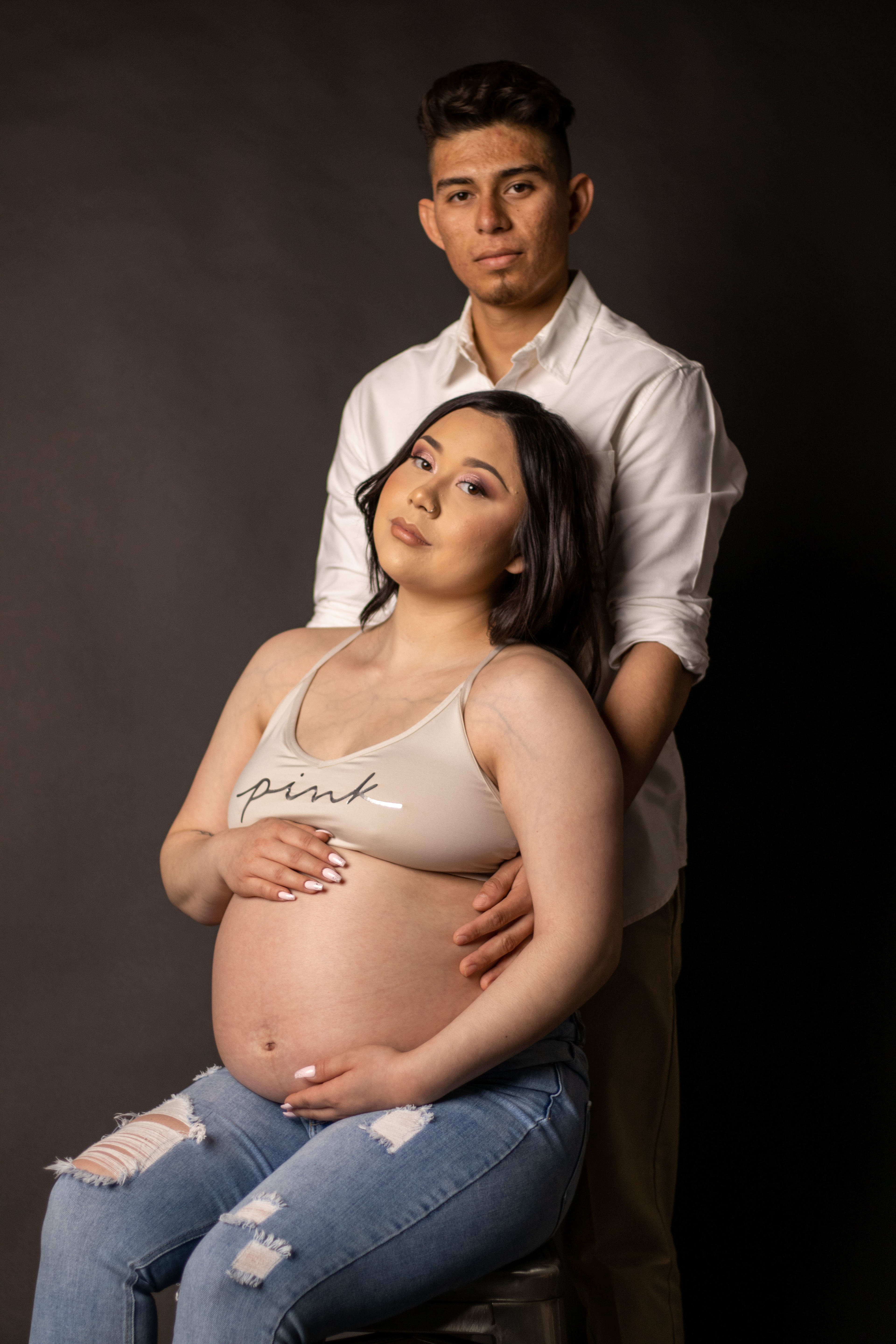 Sesión de fotos de maternidad de una mujer embarazada en un taburete con un hombre sujetándola por detrás.