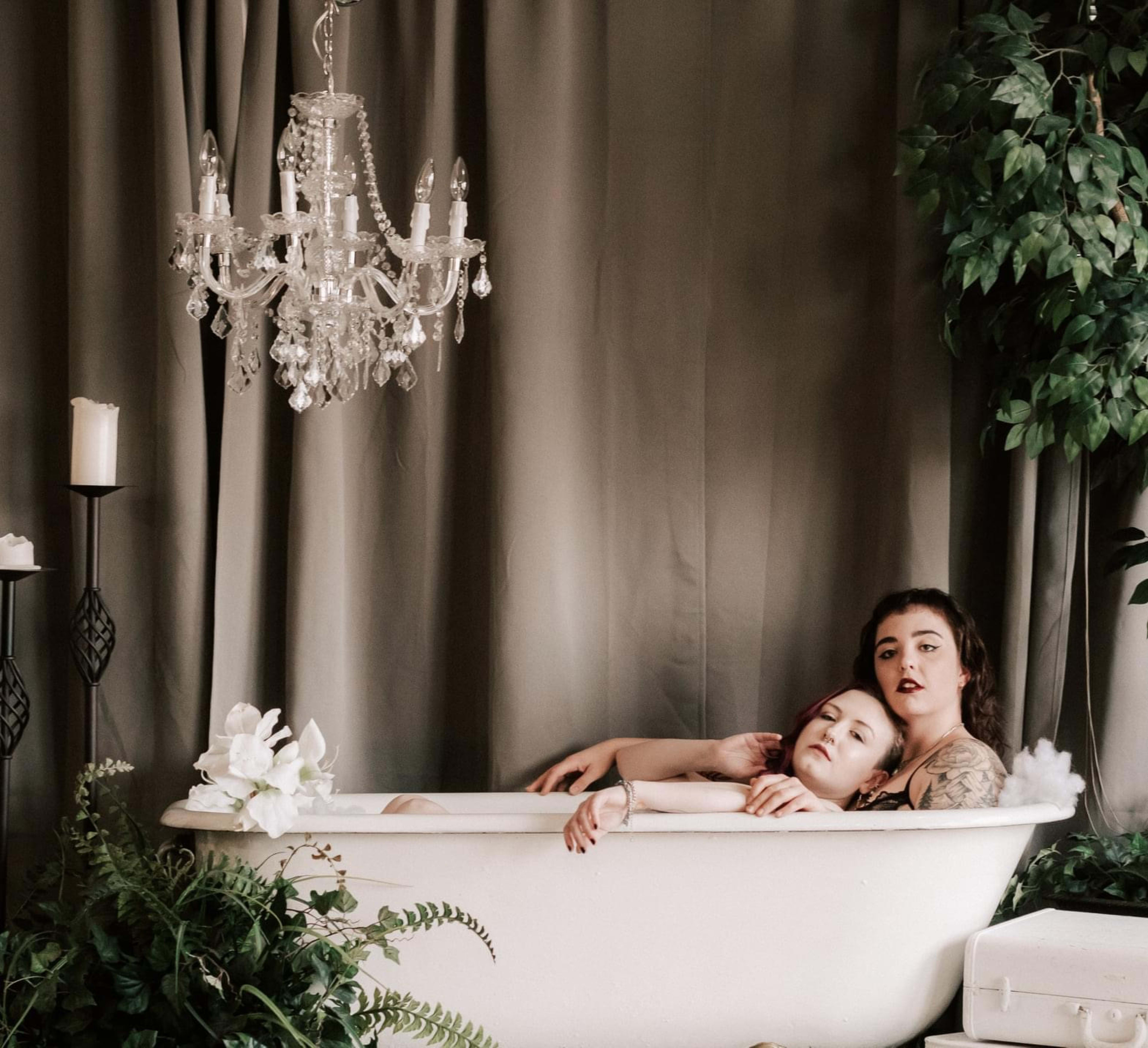 Two women in lingerie pose for a photoshoot in a bathtub surrounded by plants and a chandelier.