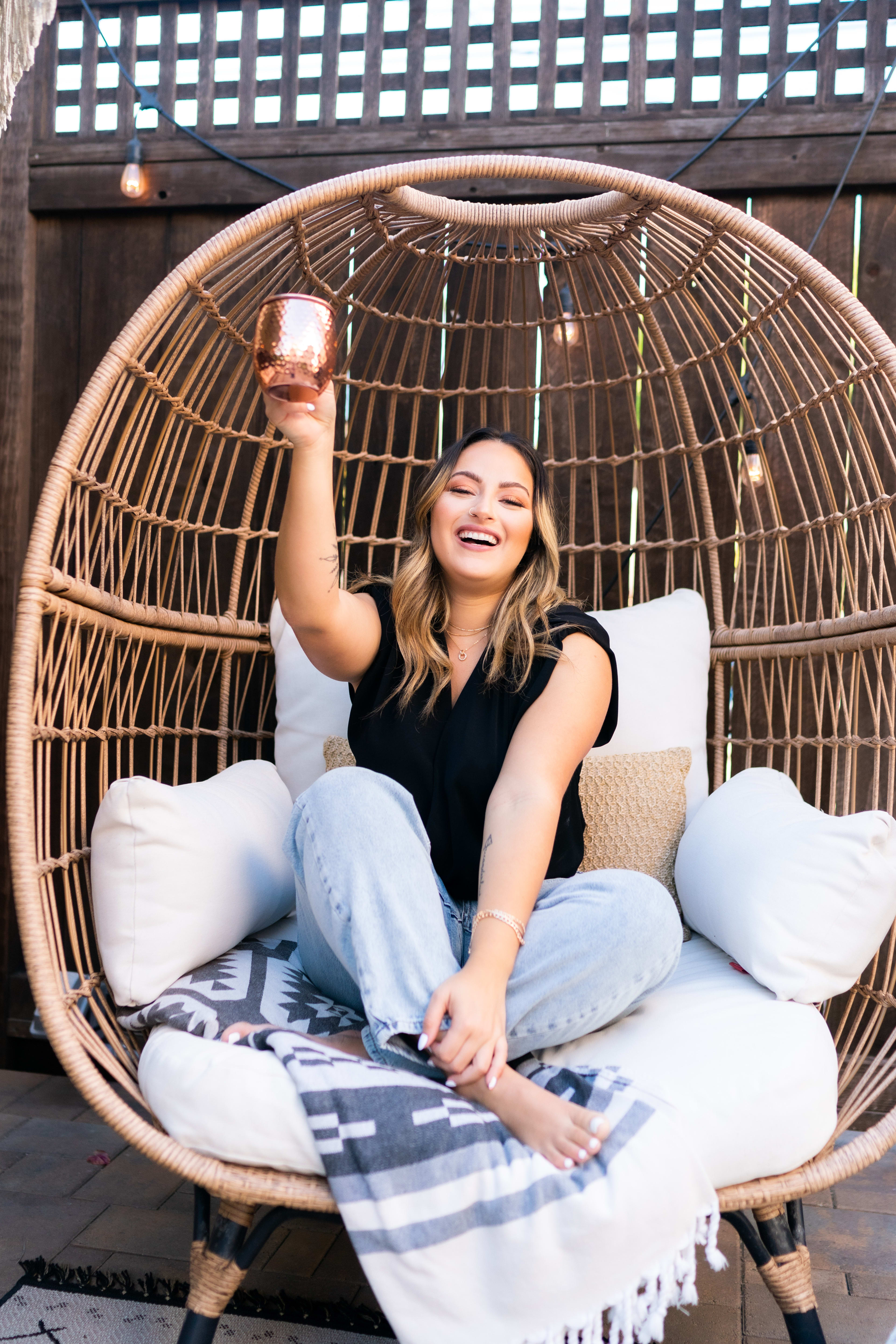 An outdoor photoshoot featuring a woman sitting in an egg chair holding a drink.