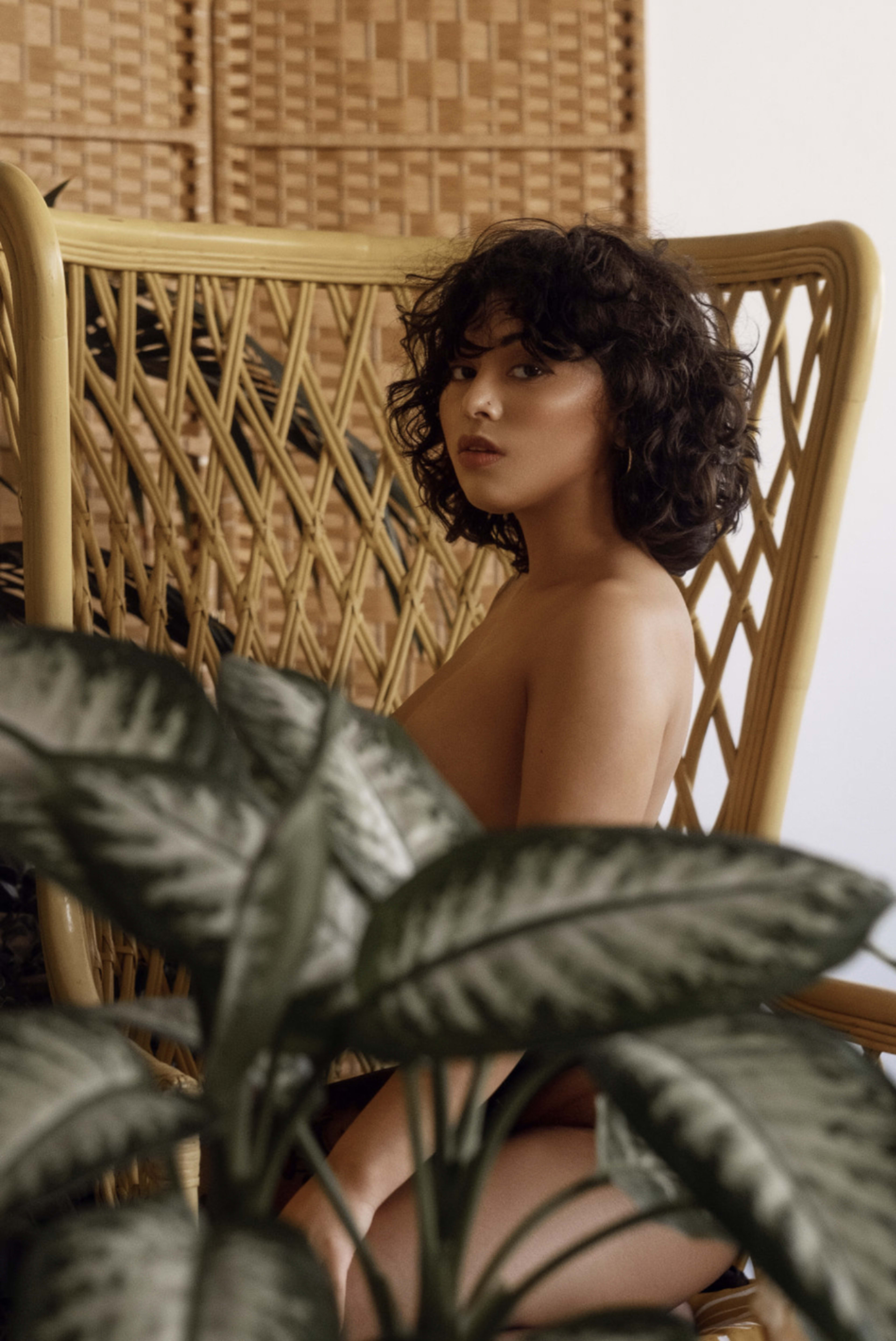 A boho-inspired boudoir photo shoot featuring a naked woman sitting in a chair surrounded by plants in shades of green and brown.