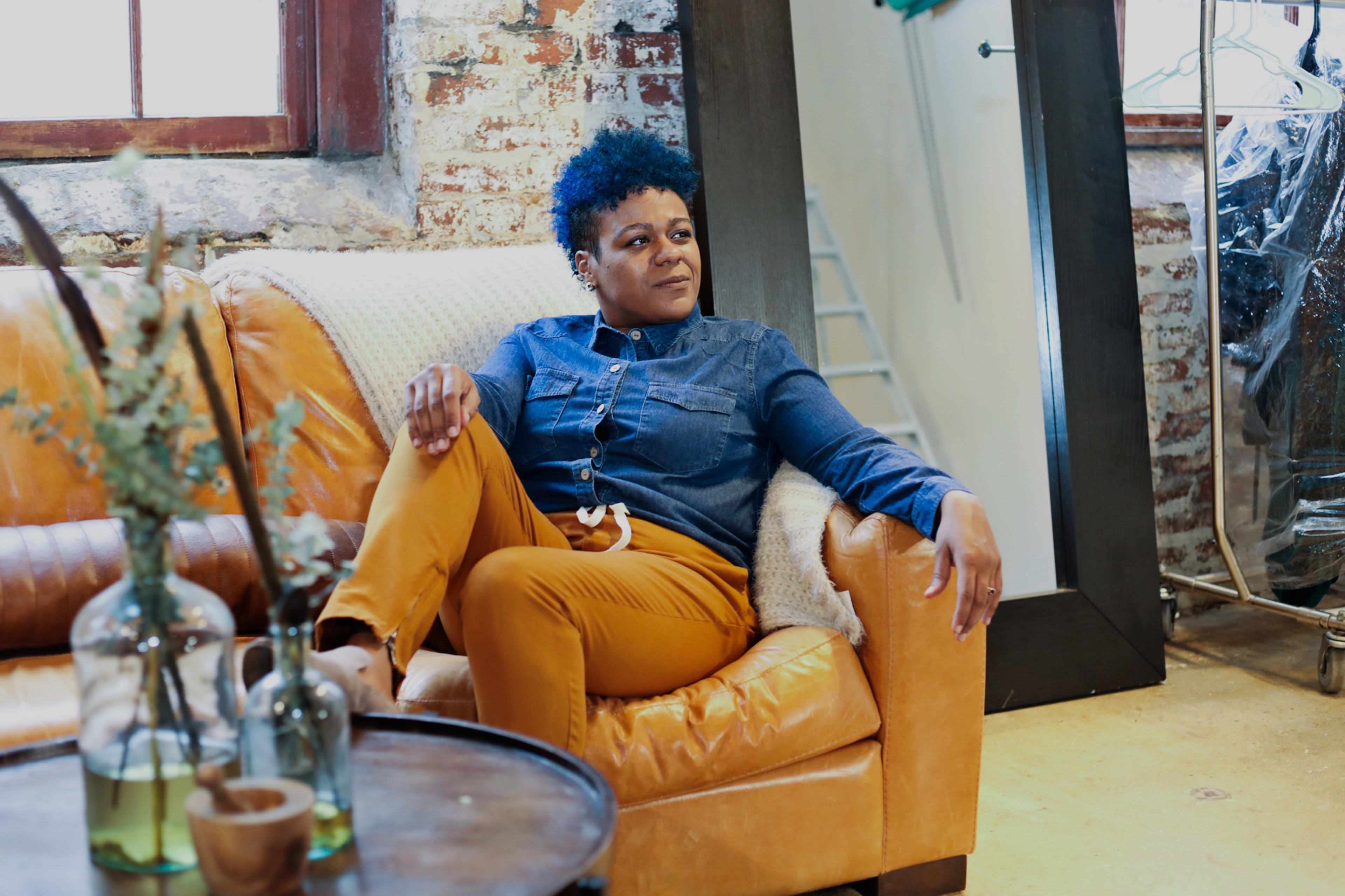 A fashion photoshoot with a woman sitting on a rustic orange couch in a room.