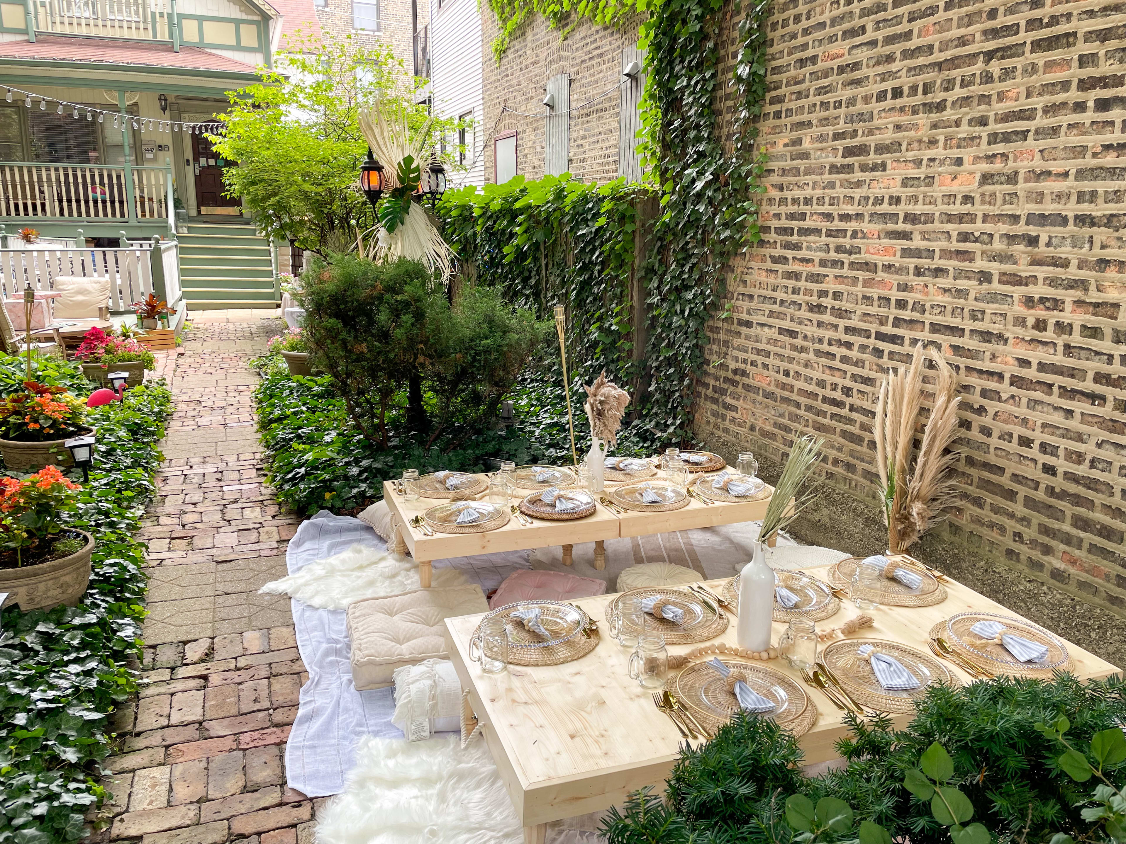 Une table de style boho est installée à l'extérieur parmi les plantes vertes d'un jardin et un mur de briques.