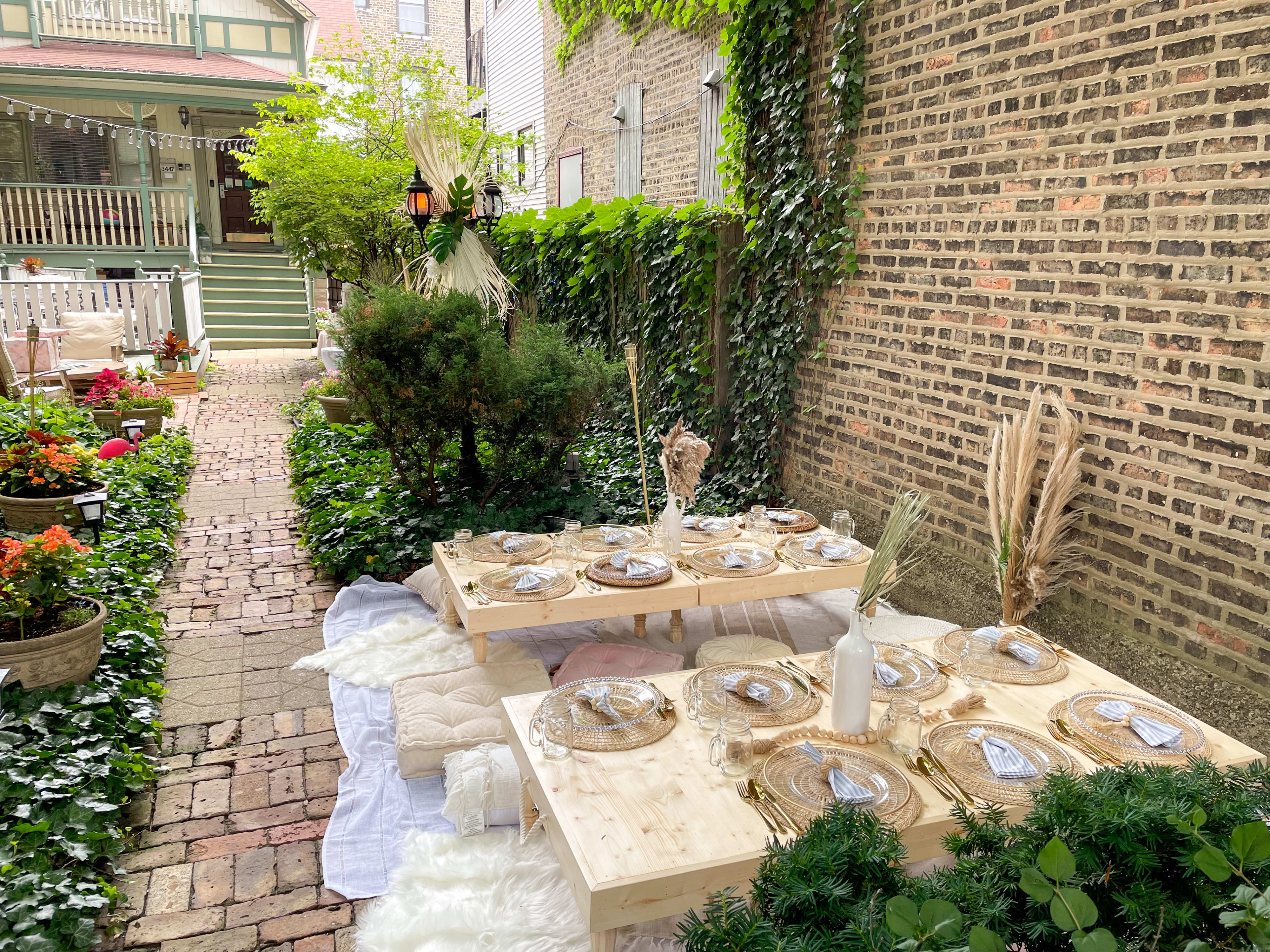 A boho table is set up outside among green plants in a garden and a brick wall.