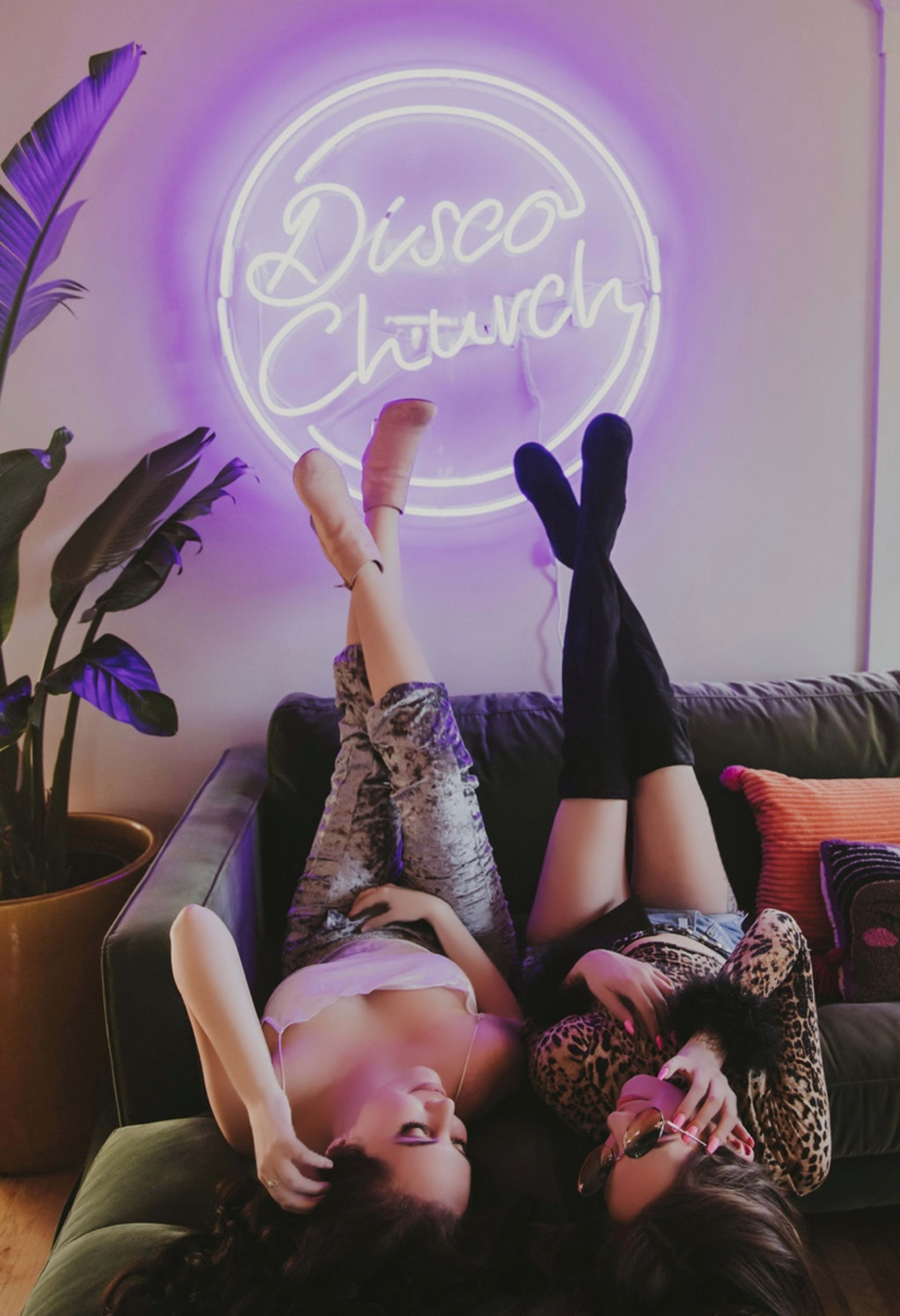 Two fashion women posing on a couch in front of a purple neon sign with plants for a photo shoot.