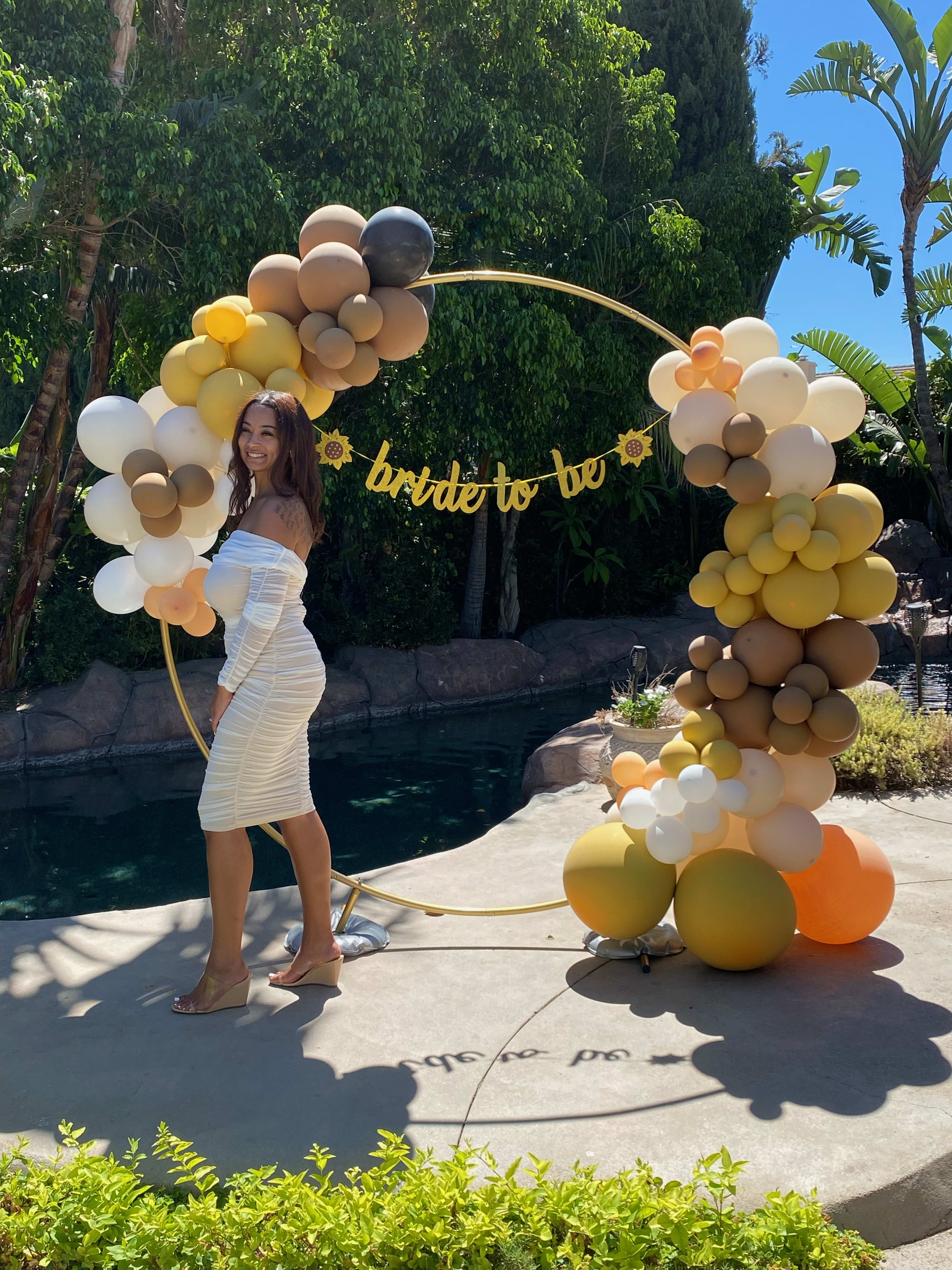 A woman in front of a bunch of yellow balloons theme arc in an outdoor garden in a bridal shower.