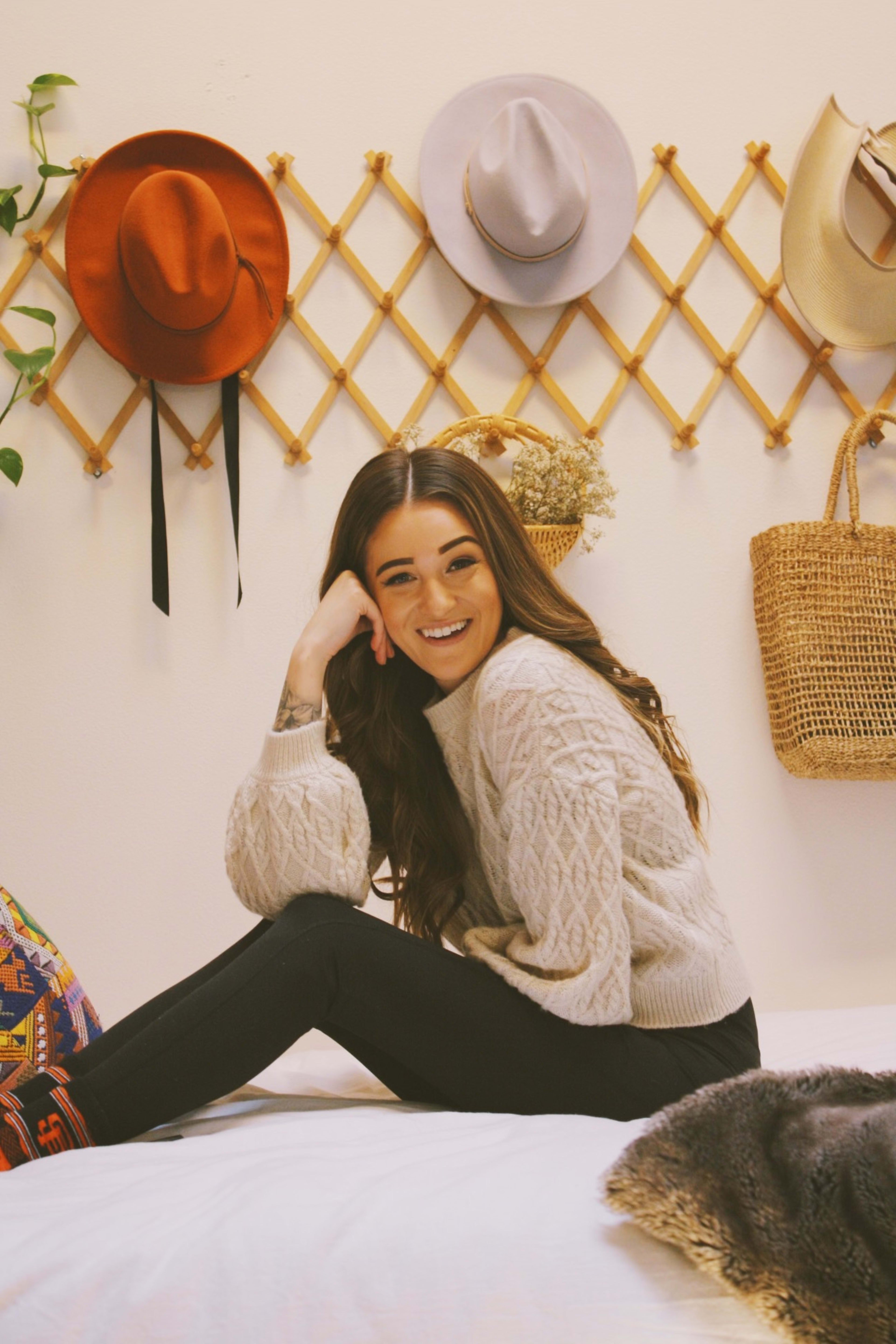 A woman posing for a fashion photo shoot on a white bed with a hat rack nearby.