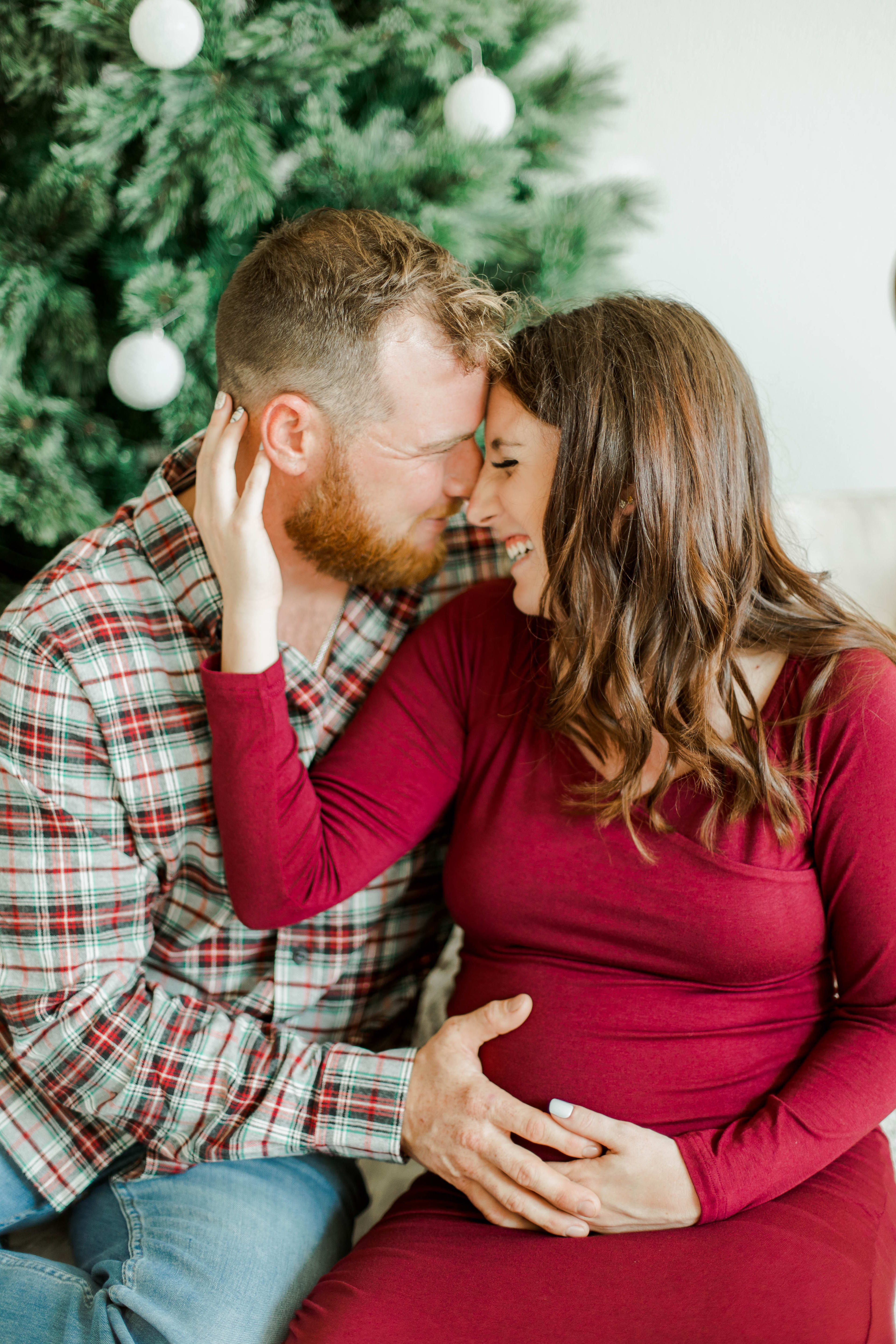 A maternity shoot of a couple cuddling in front of a Christmas tree.