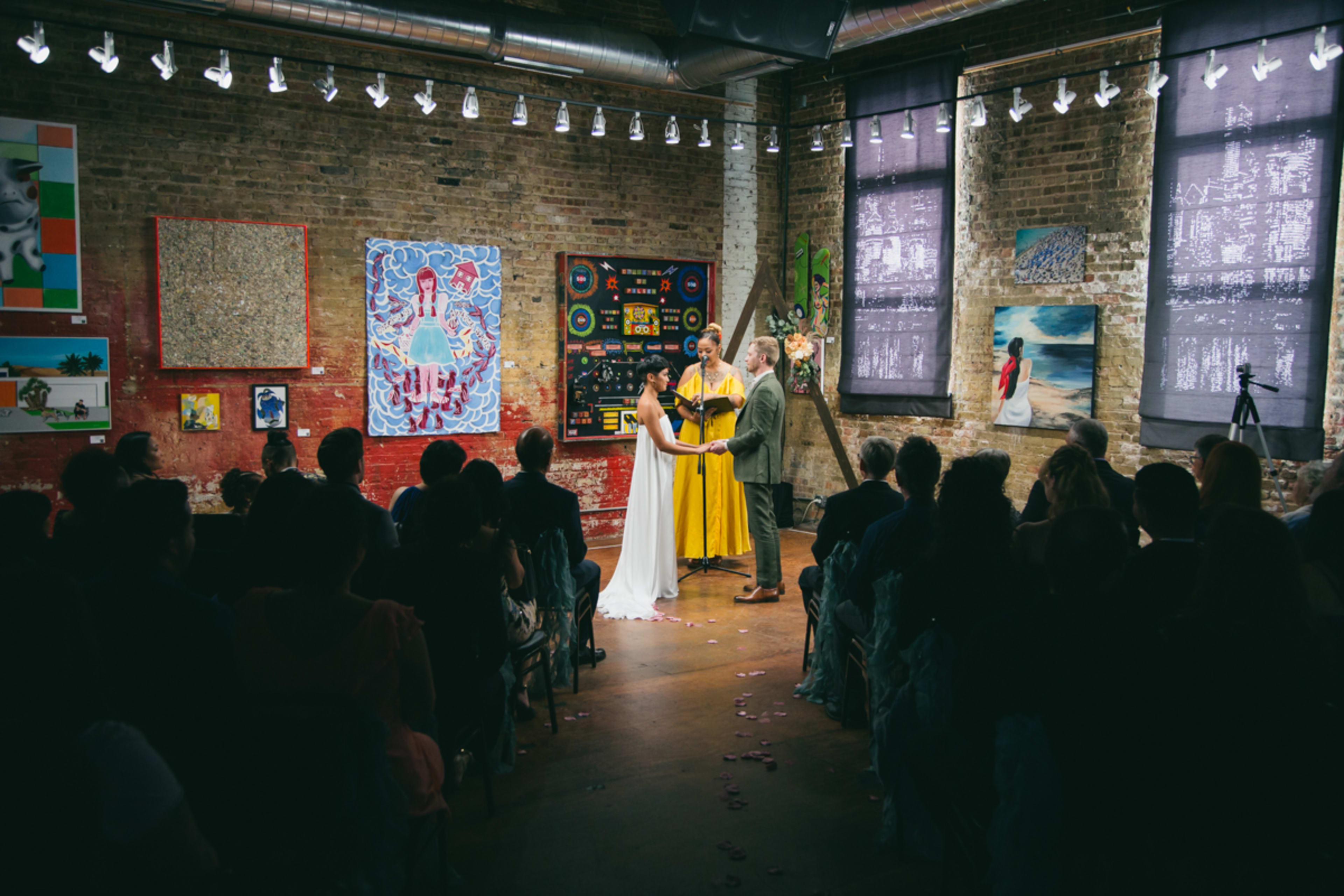 A couple getting married in front of a crowd at a wedding in an art gallery with exposed brick.