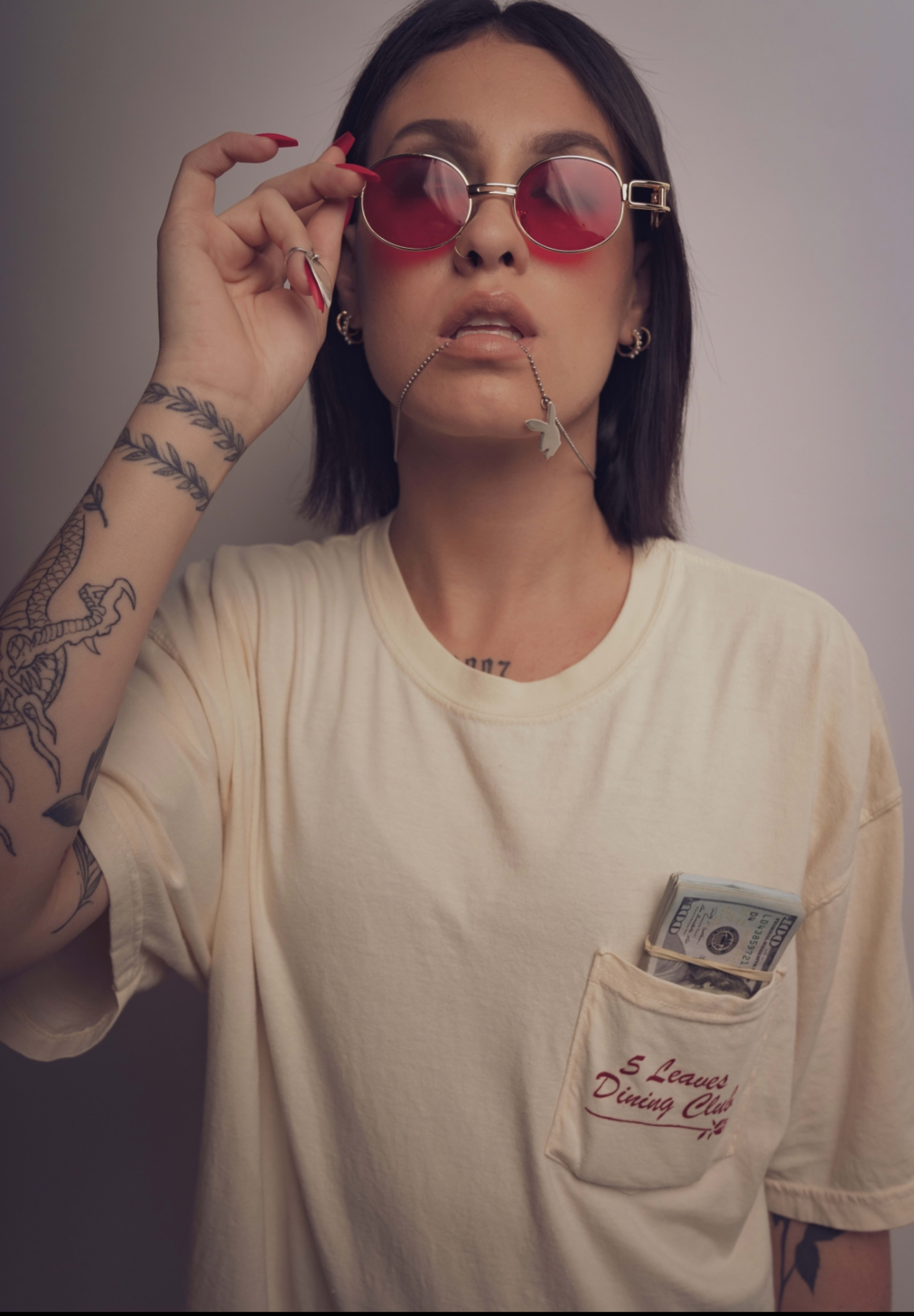 Una modelo con tatuajes, camisa blanca y gafas rojas con dinero posando para una sesión de fotos.