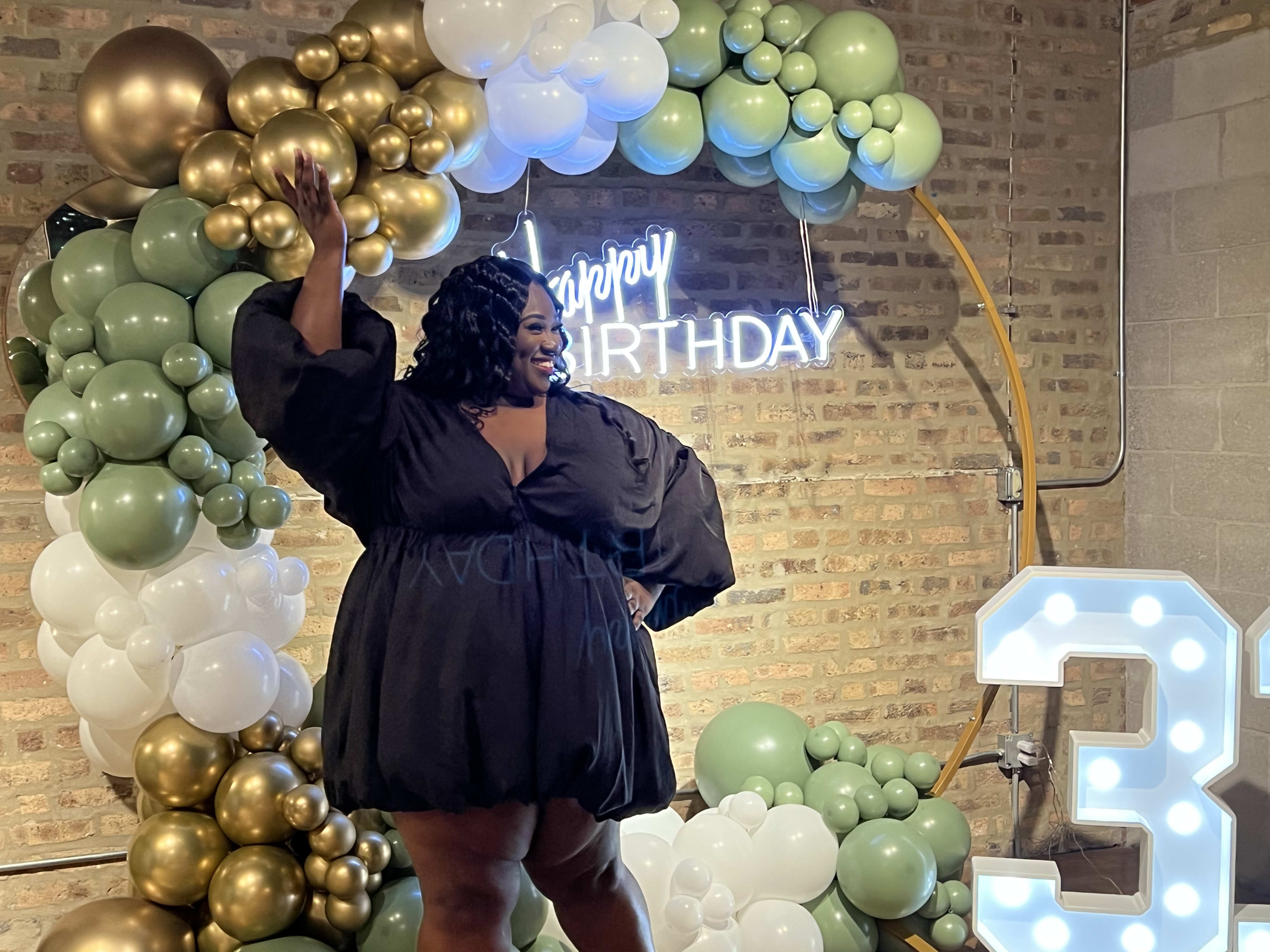 Una mujer con un vestido negro delante de globos en una fiesta de cumpleaños.