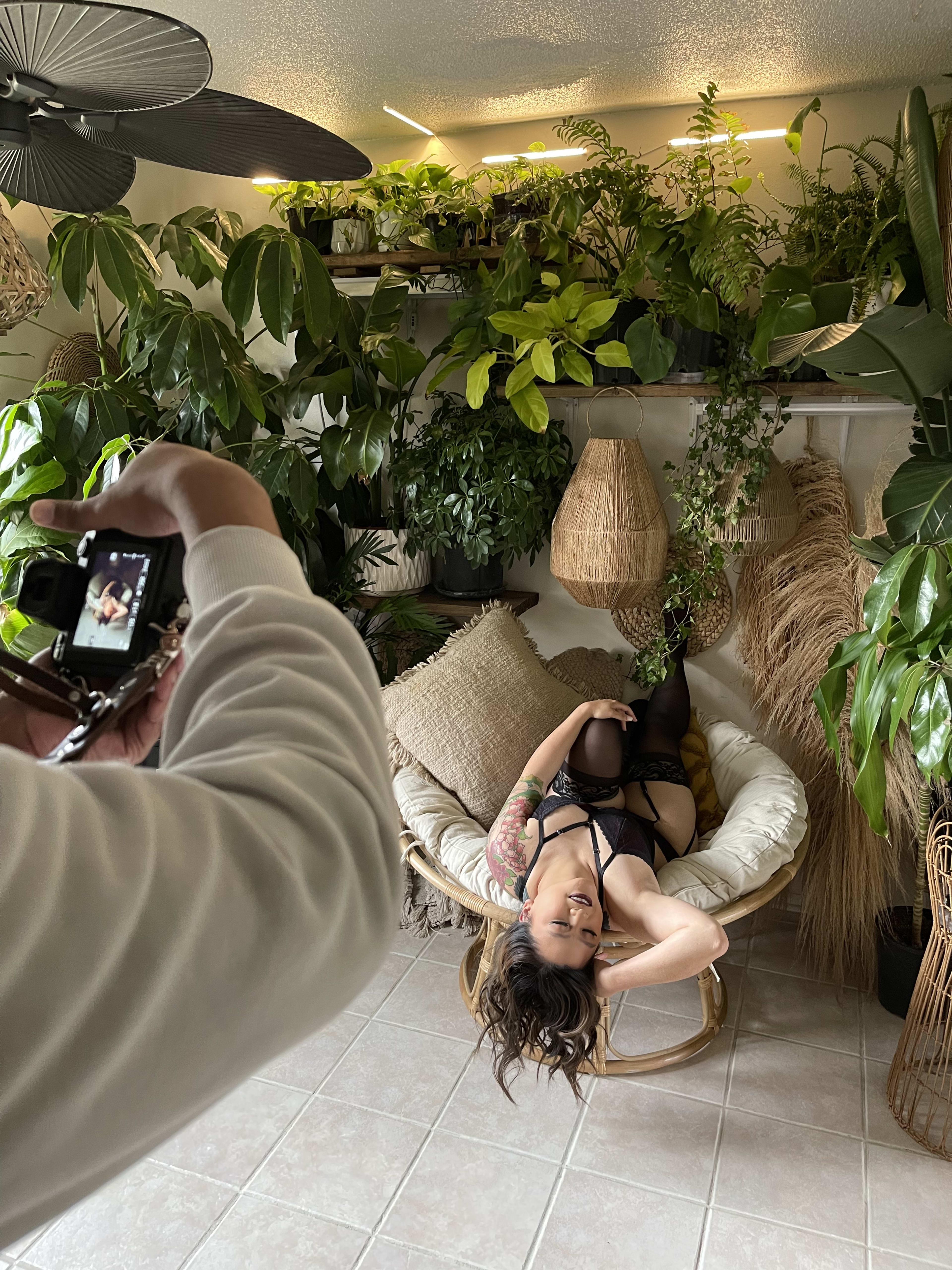 A woman is posing for a photoshoot in front of a green plants.