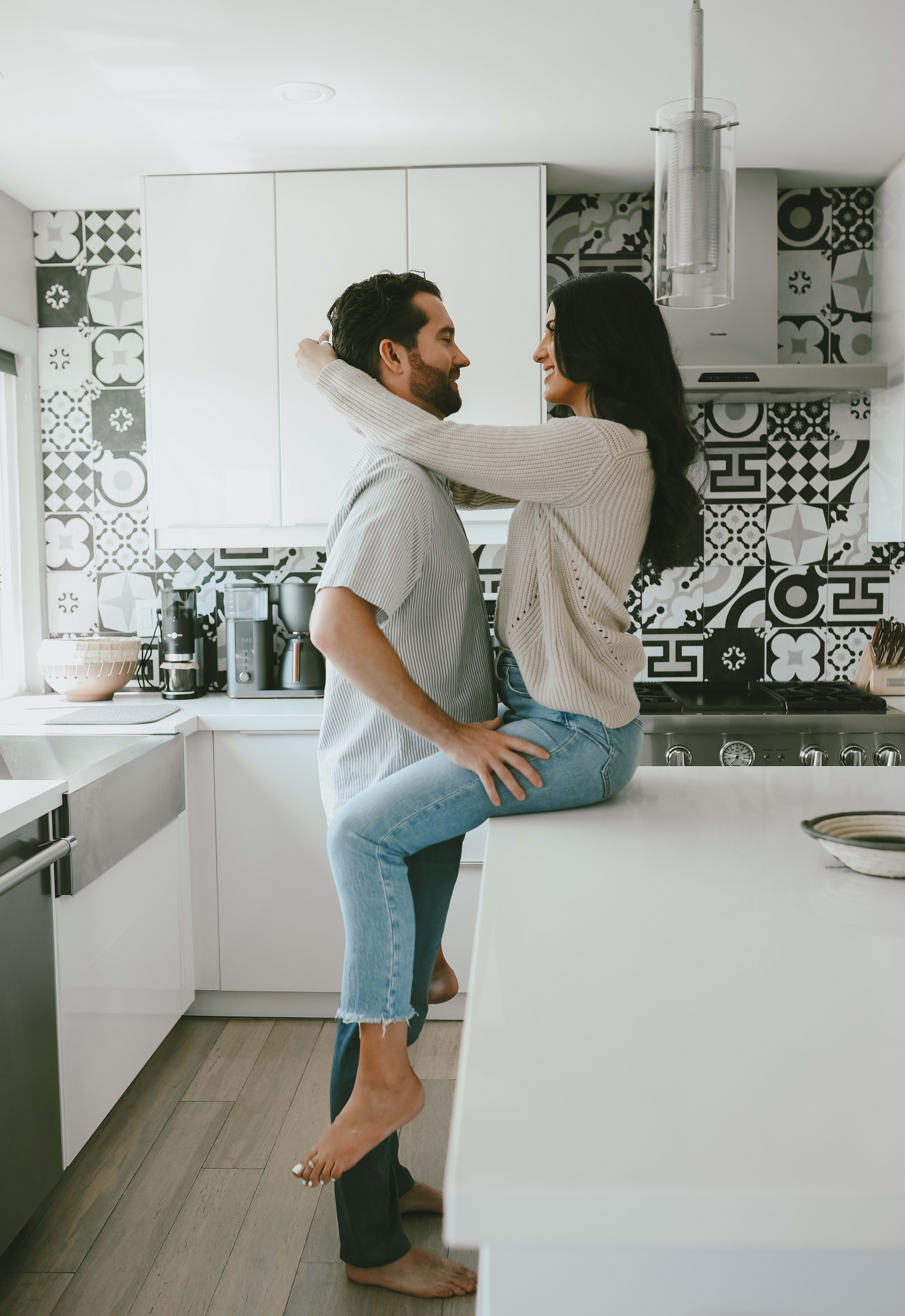 A couple poses for a photoshoot in their white kitchen, with grey accents adding subtle detail to the background.