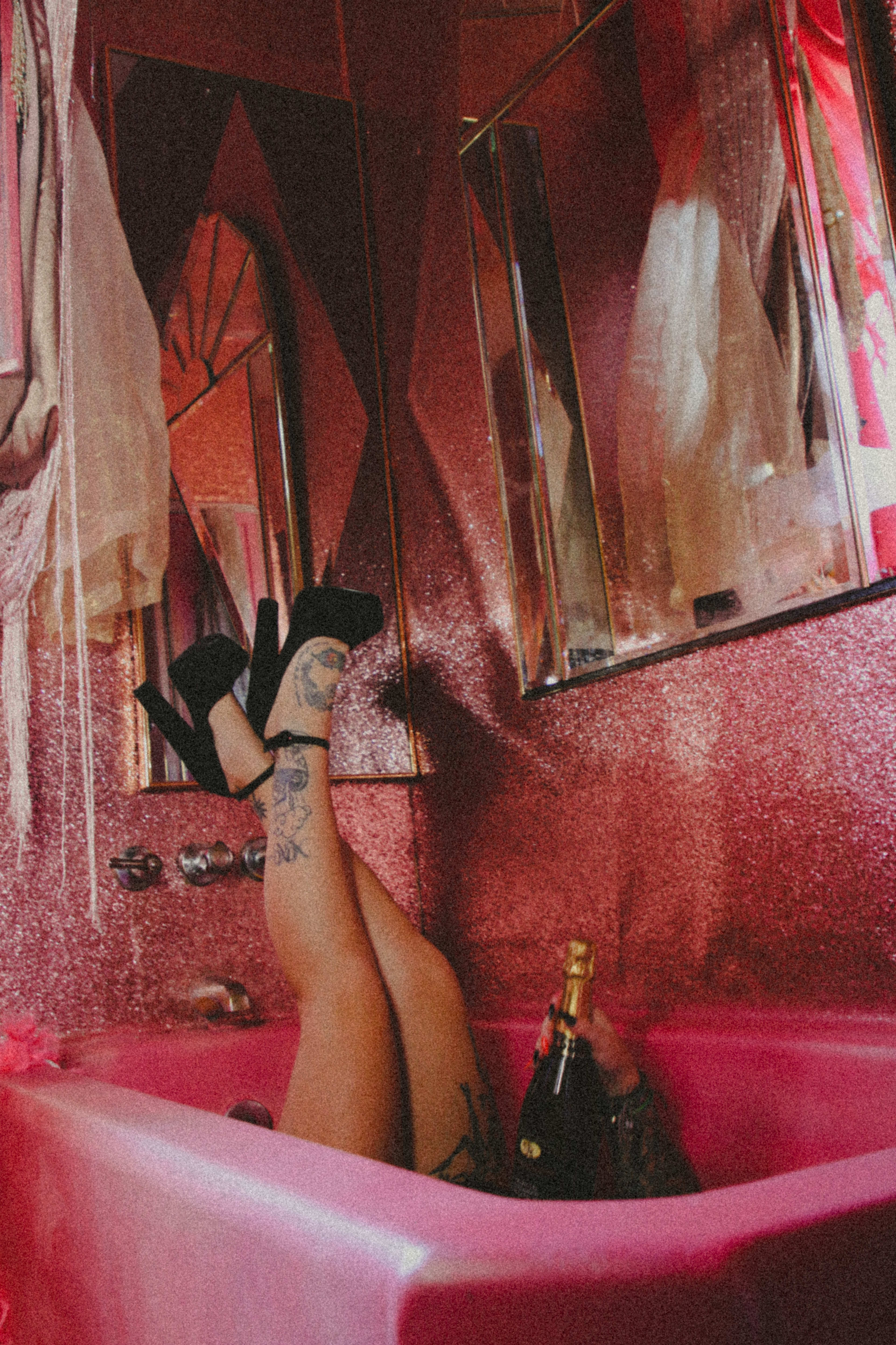 A pink photo shoot of a woman's legs in a bath with a bottle of wine.