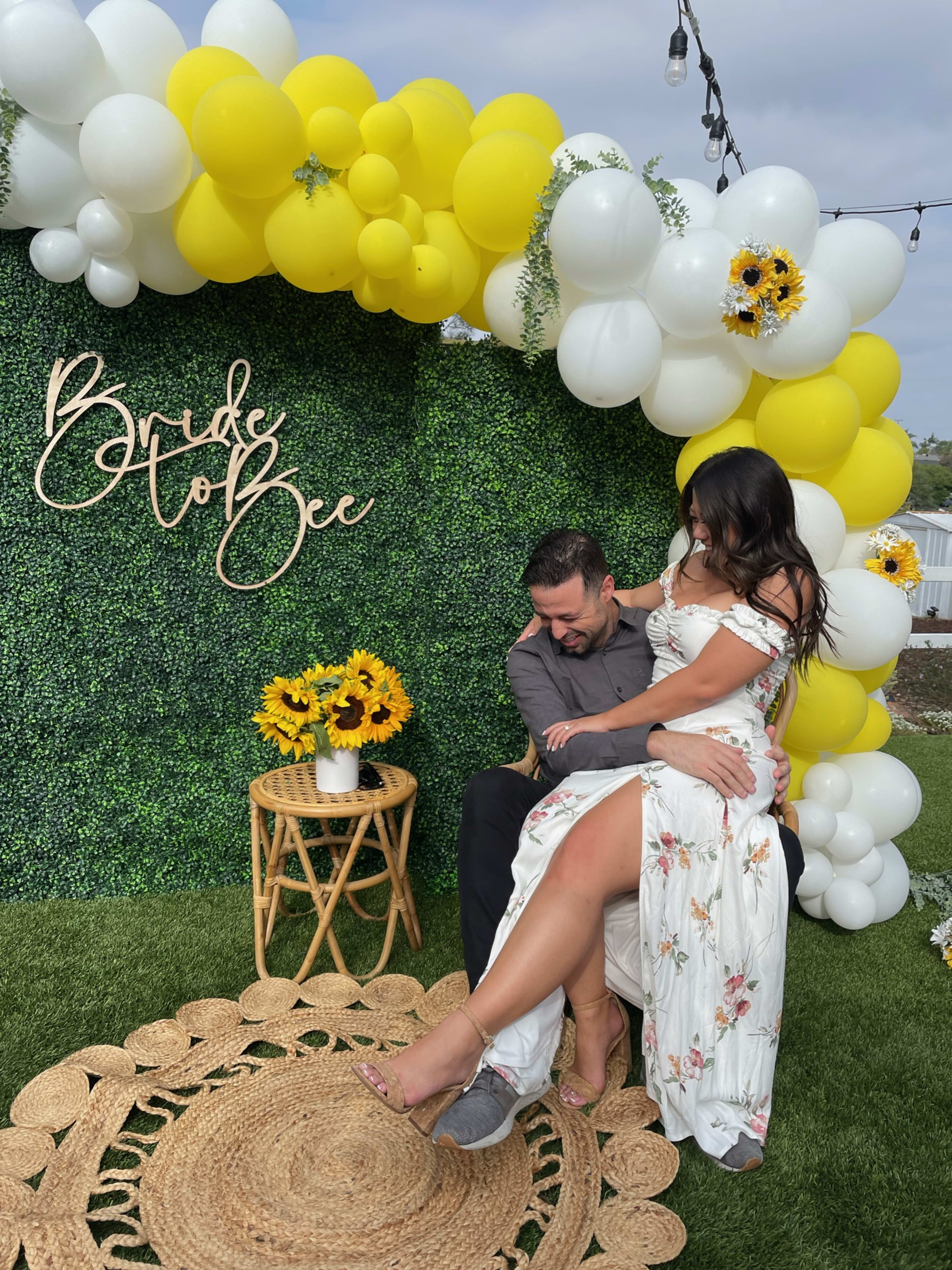 Una pareja sentada en una alfombra bajo un arco de globos boho amarillos y blancos.