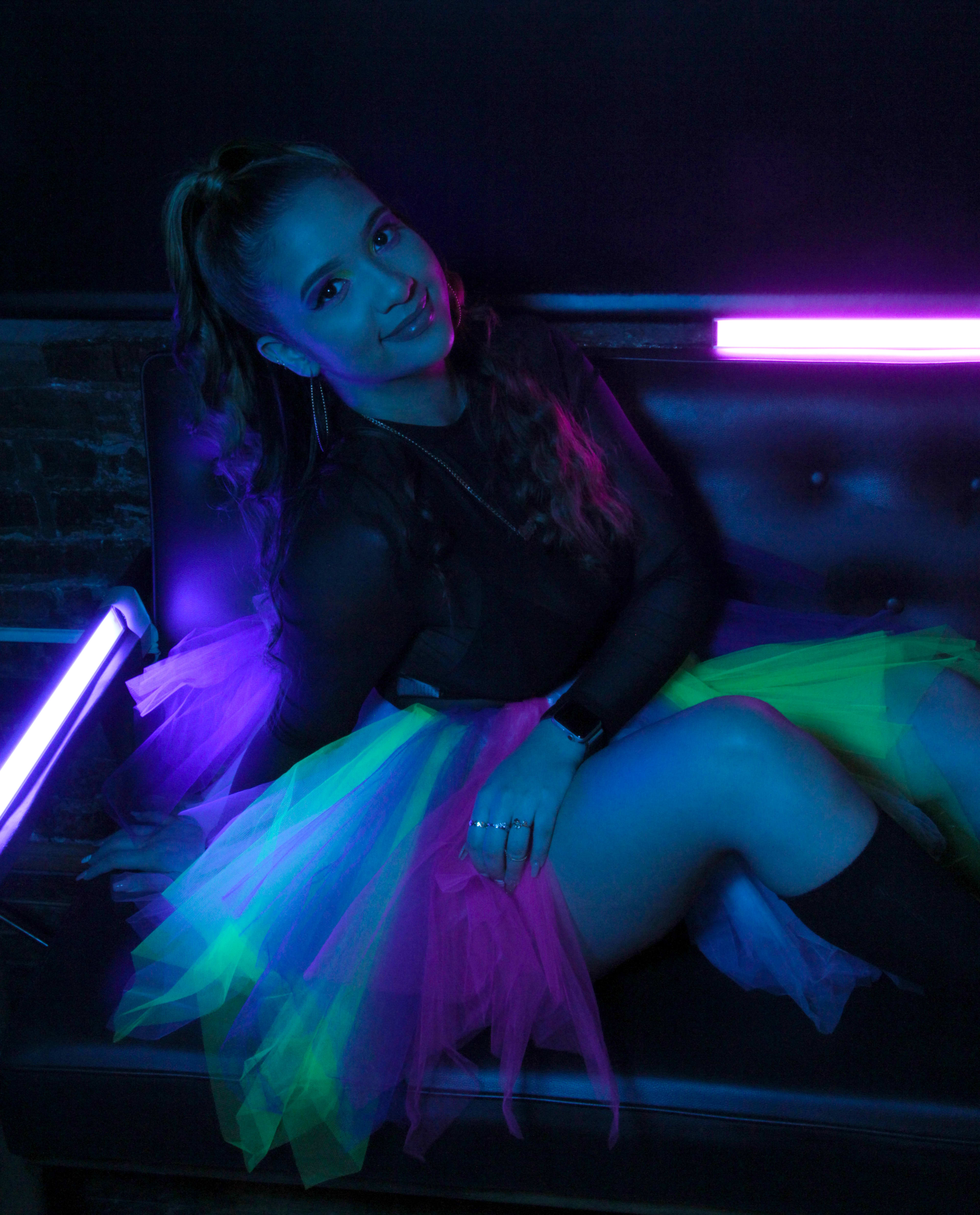 A woman posing for a photoshoot on a neon-lit couch in an 1980s inspired fashion setting.
