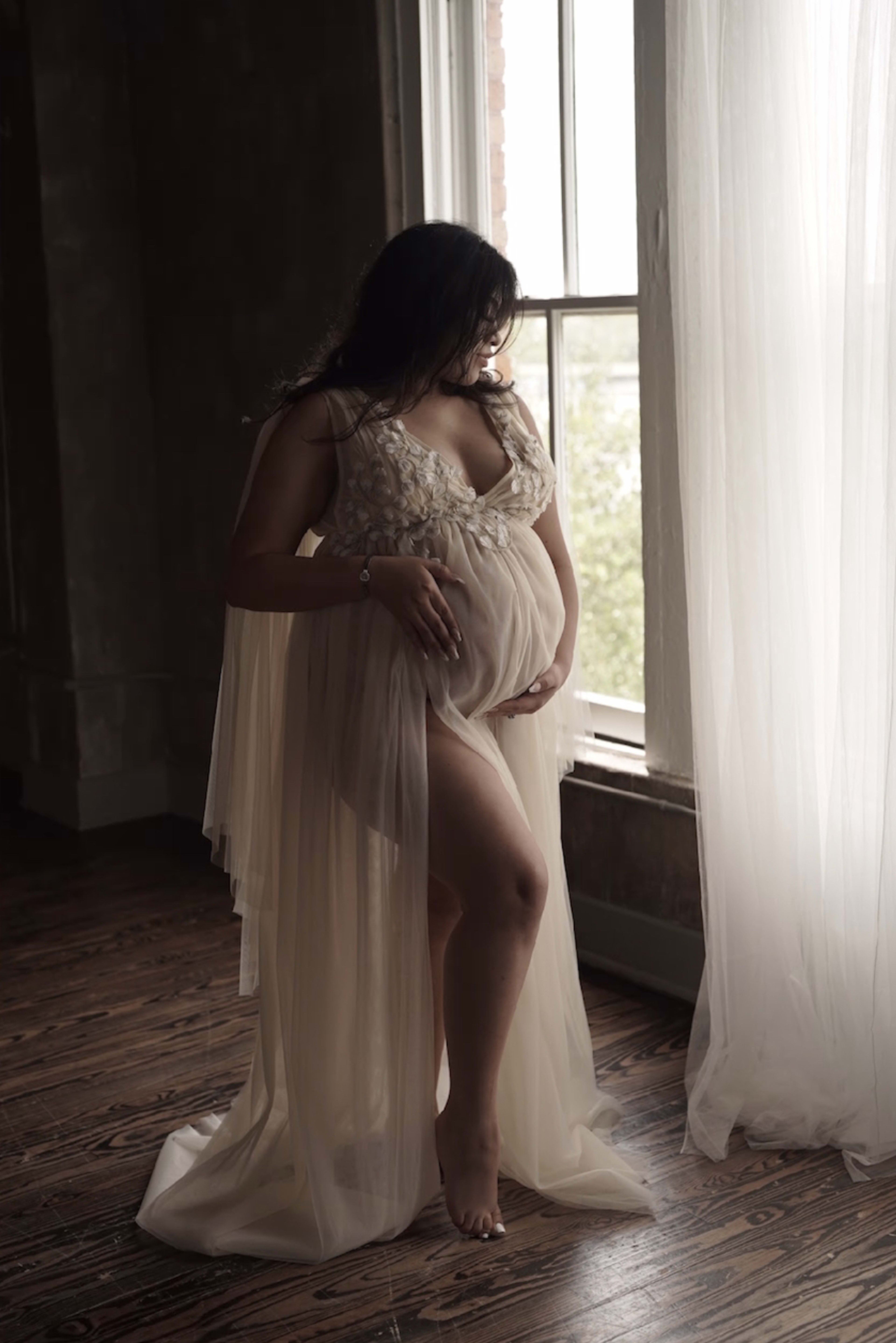 A maternity photo shoot of a woman in a white dress standing in front of a window.