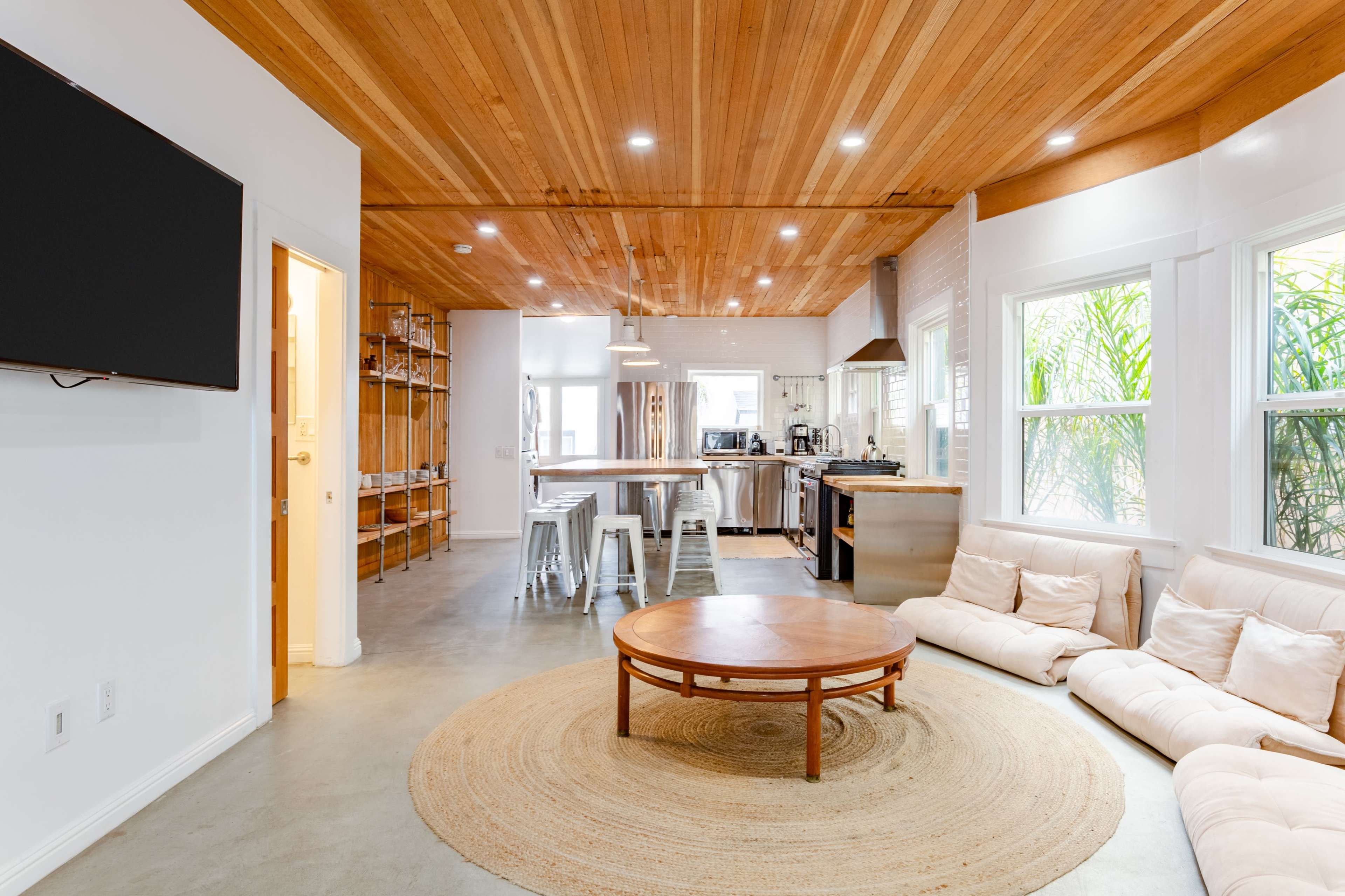 A modern living room with a wooden ceiling, a round coffee table on a woven rug, and a sectional sofa facing a wall-mounted television, with an open kitchen visible in the background.