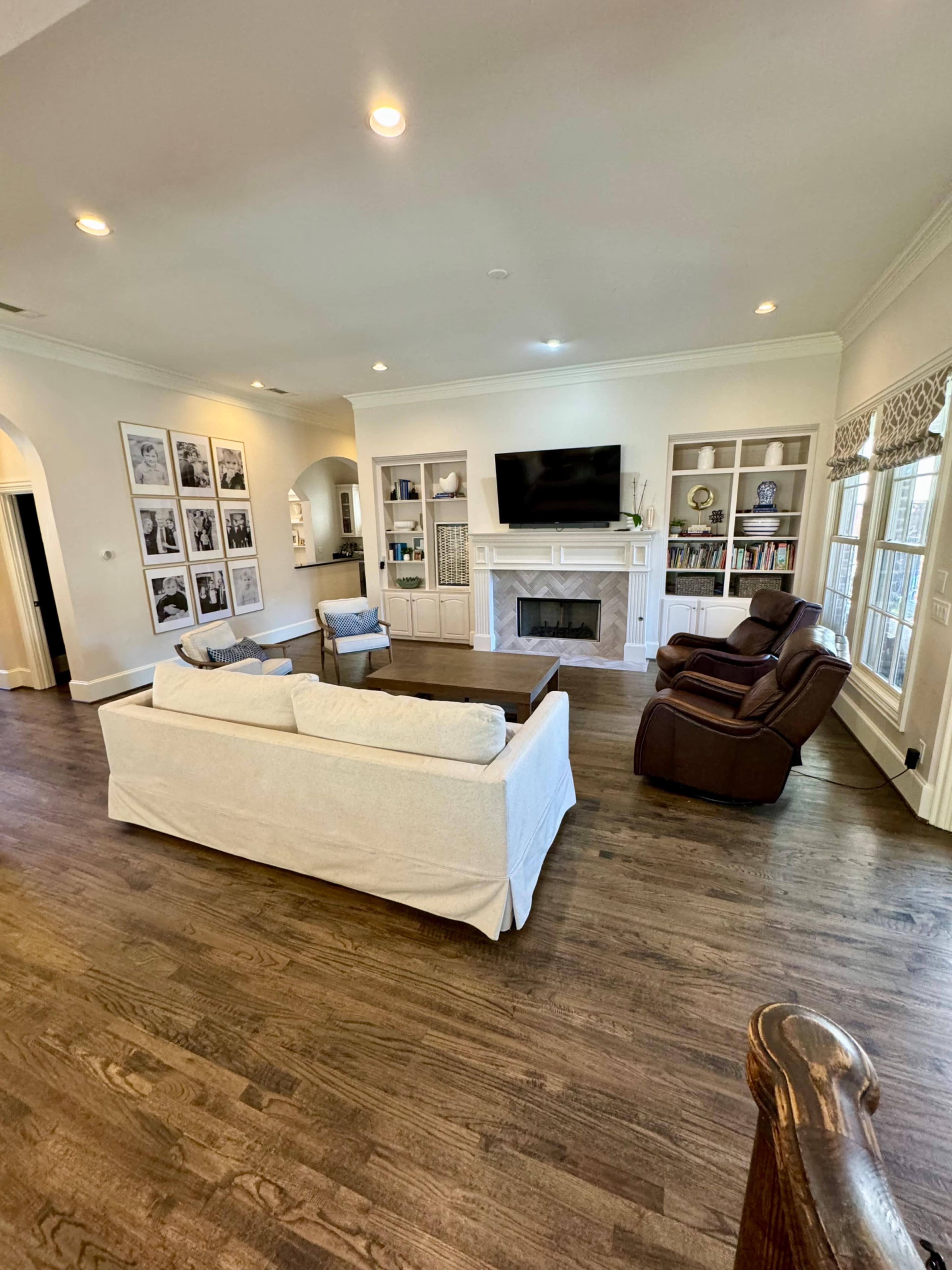 A spacious living room features a light-colored sofa, two brown armchairs, a large coffee table, and built-in shelves on either side of a television above a fireplace.