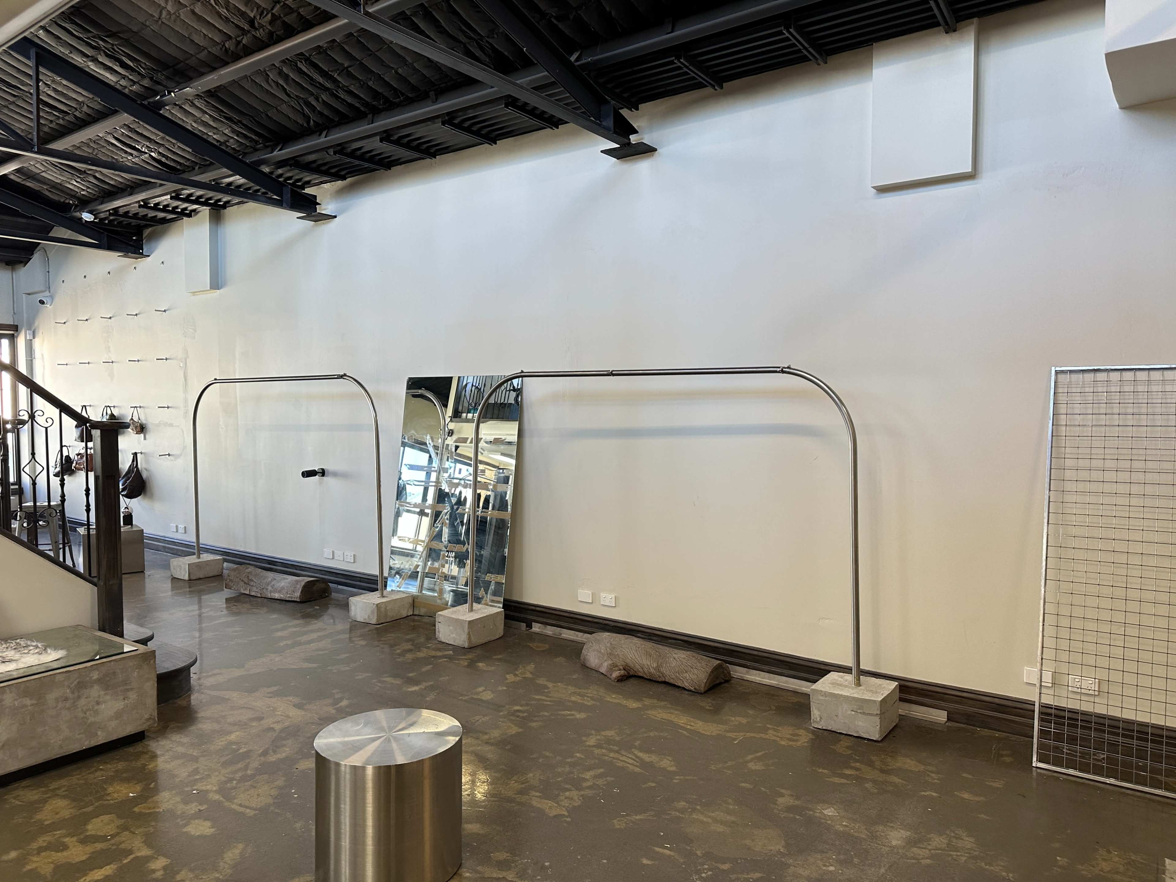 The image shows a minimalist interior space with a large standing mirror, metal frames, and exposed concrete elements against a plain wall.