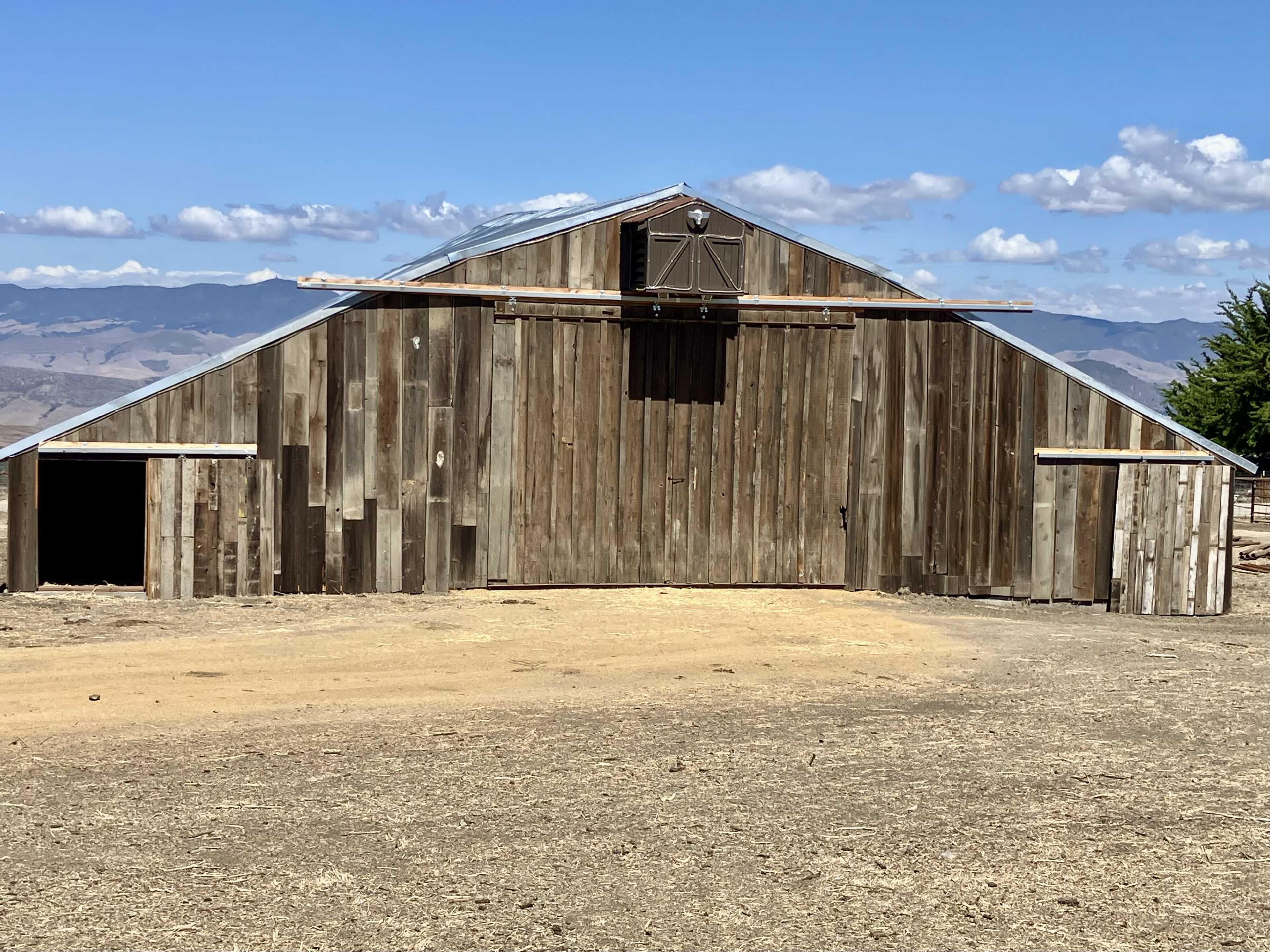 A large wooden barn with a sloped roof sits on a dirt lot, surrounded by hills and blue skies.