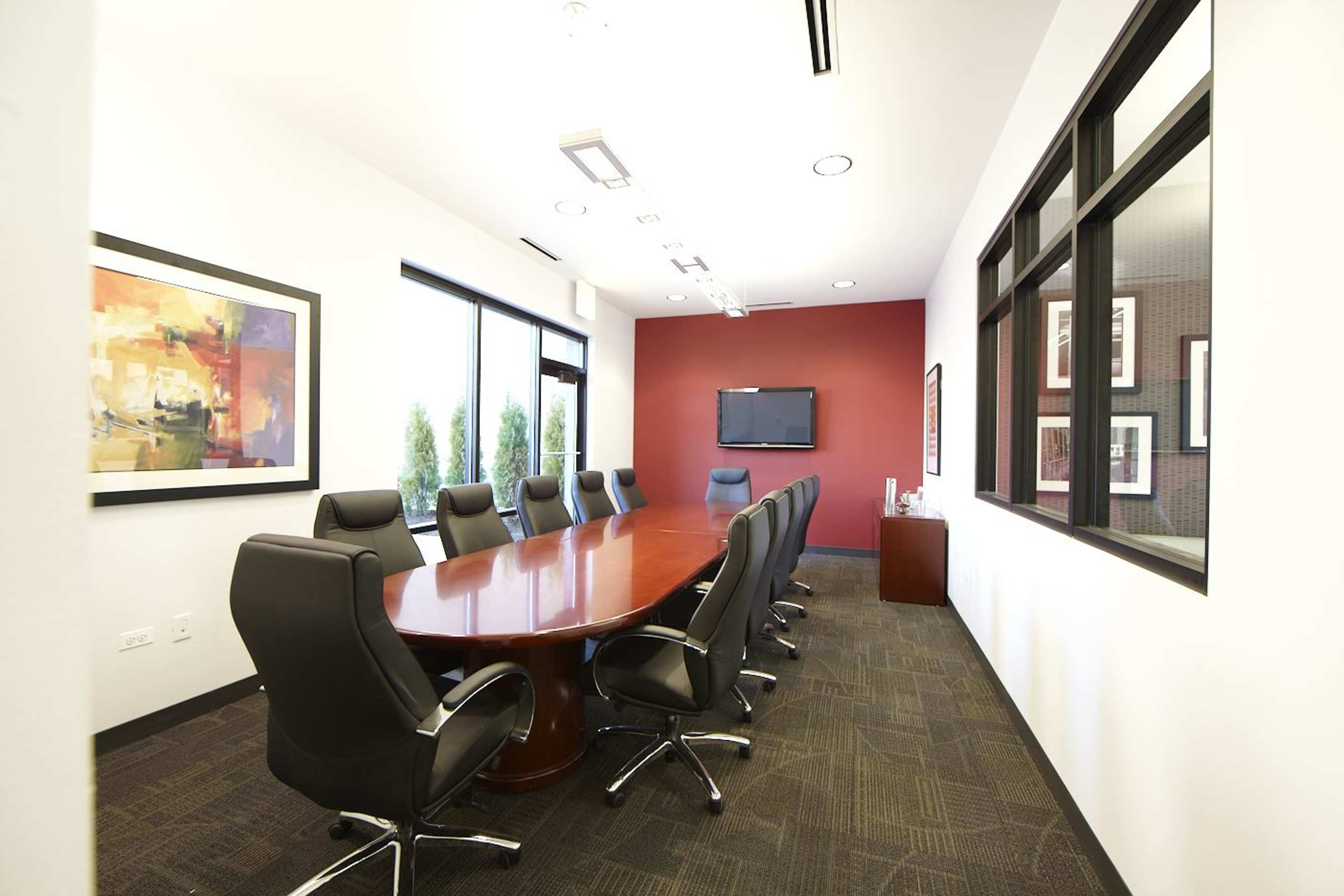 A spacious conference room features a large wooden table surrounded by black leather chairs and is equipped with a wall-mounted television and artwork.