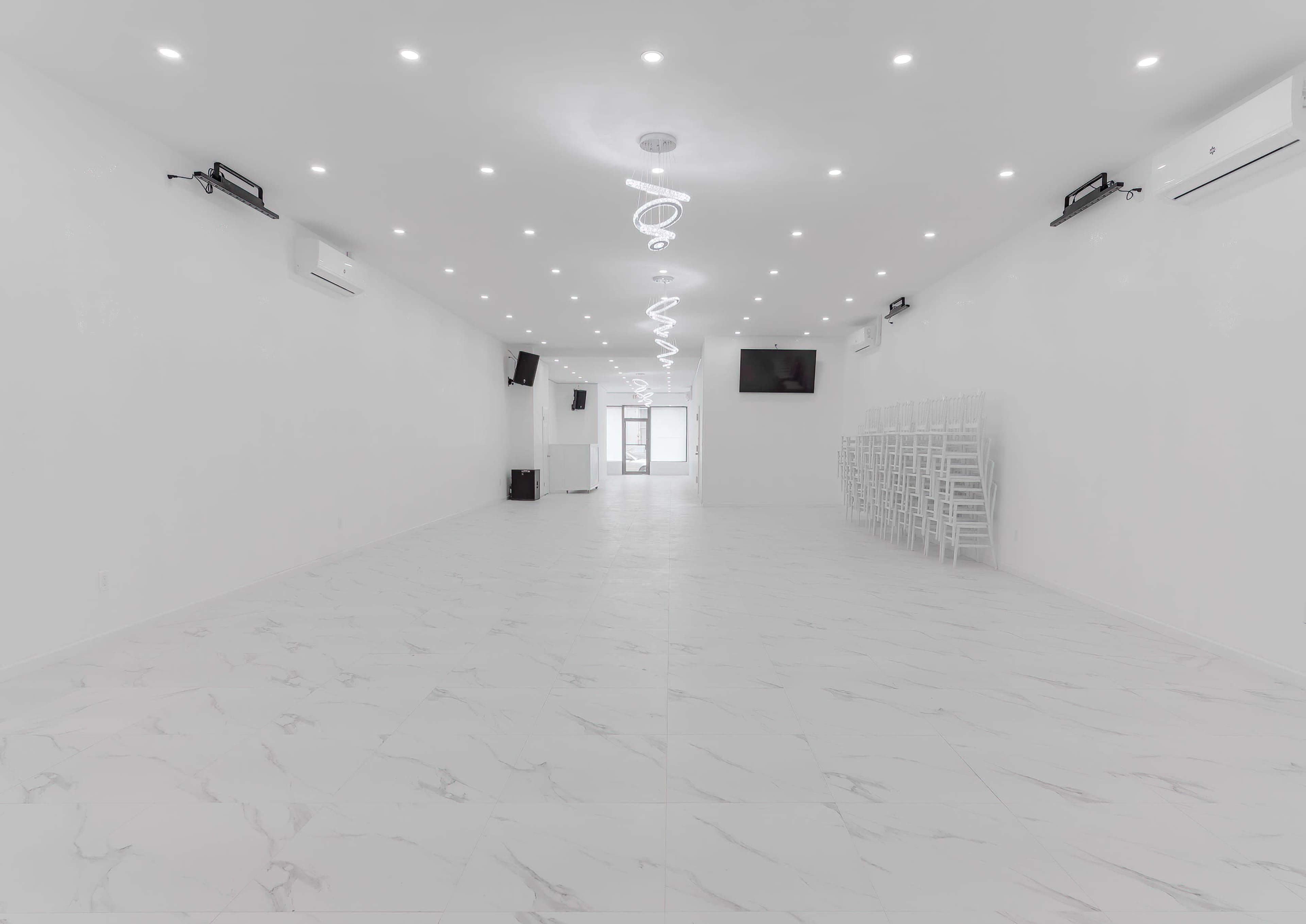 The interior of a spacious, bright room features white walls, marble-patterned flooring, and rows of stackable chairs against one side.