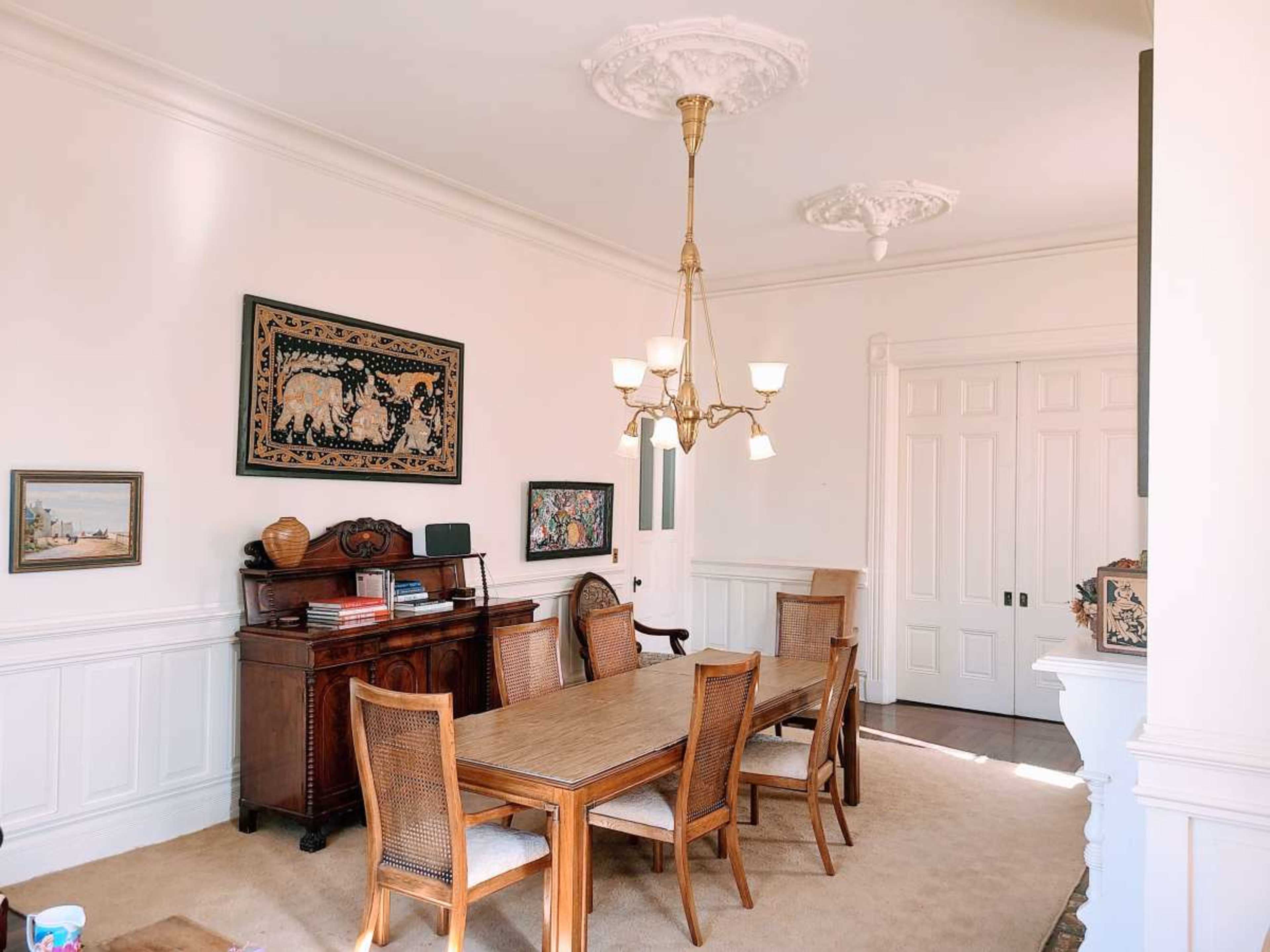 The image shows a spacious dining room featuring a wooden table surrounded by chairs, with decorative wall art and a vintage sideboard against the wall.