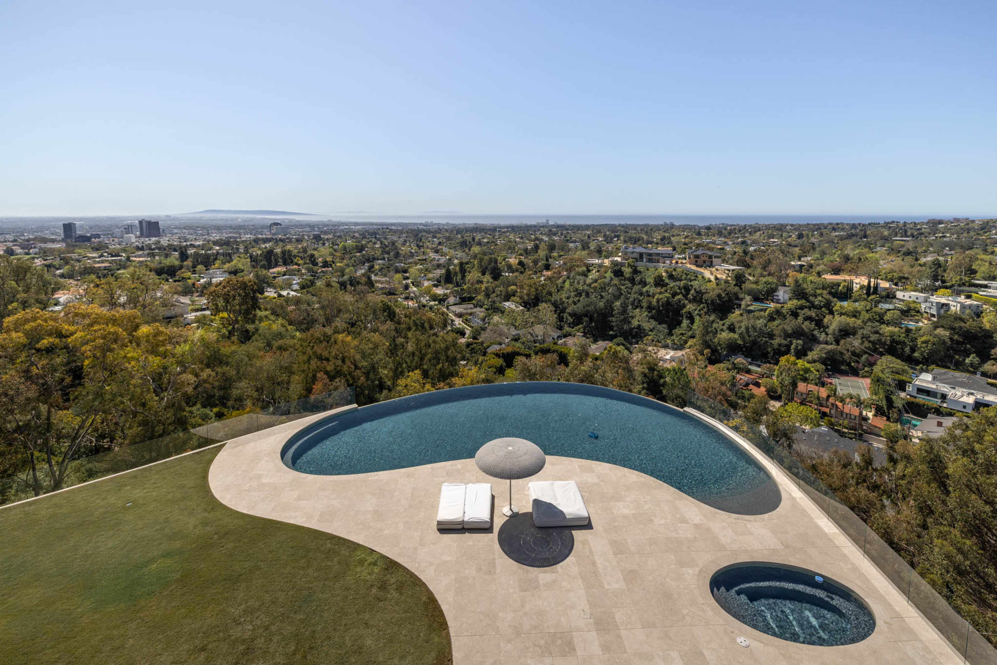 The image shows a modern infinity pool with a sunbathing area and a round table set against a panoramic view of a city and surrounding landscape.