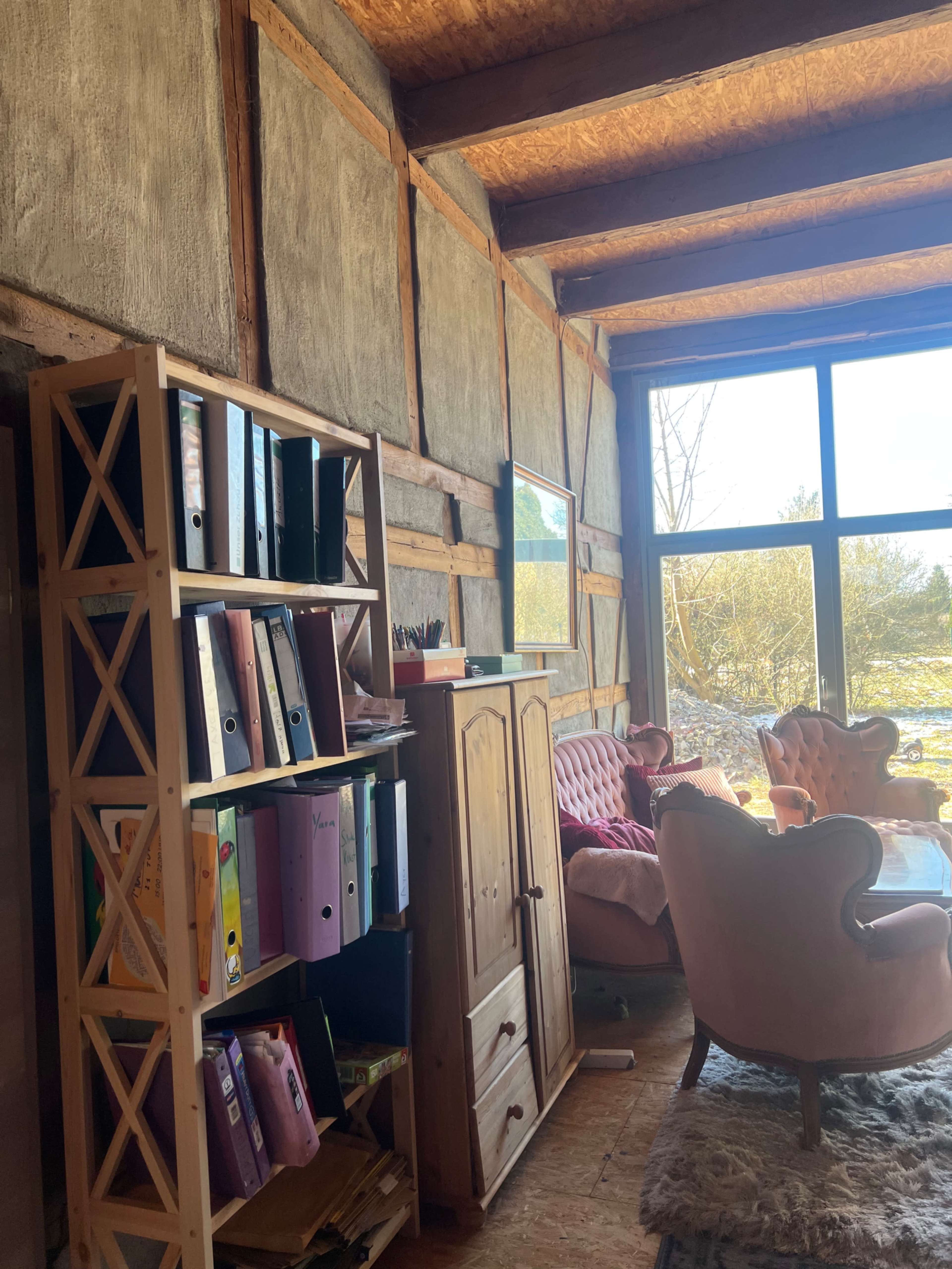 The image shows a cozy room with wooden beams, a bookshelf filled with binders, and two pink armchairs by a large window overlooking a natural landscape.
